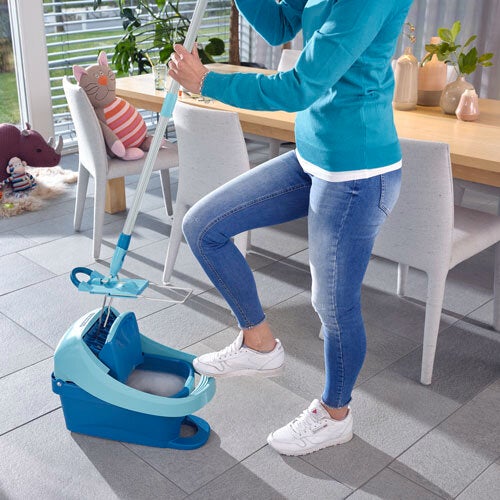 Eine Person reinigt den Boden mit einem modernen Wischsystem. Der Wischer hat einen blauen Eimer mit integriertem Auswringsystem, ideal für effiziente Reinigung. Im Hintergrund sind ein Tisch und Stühle zu sehen.