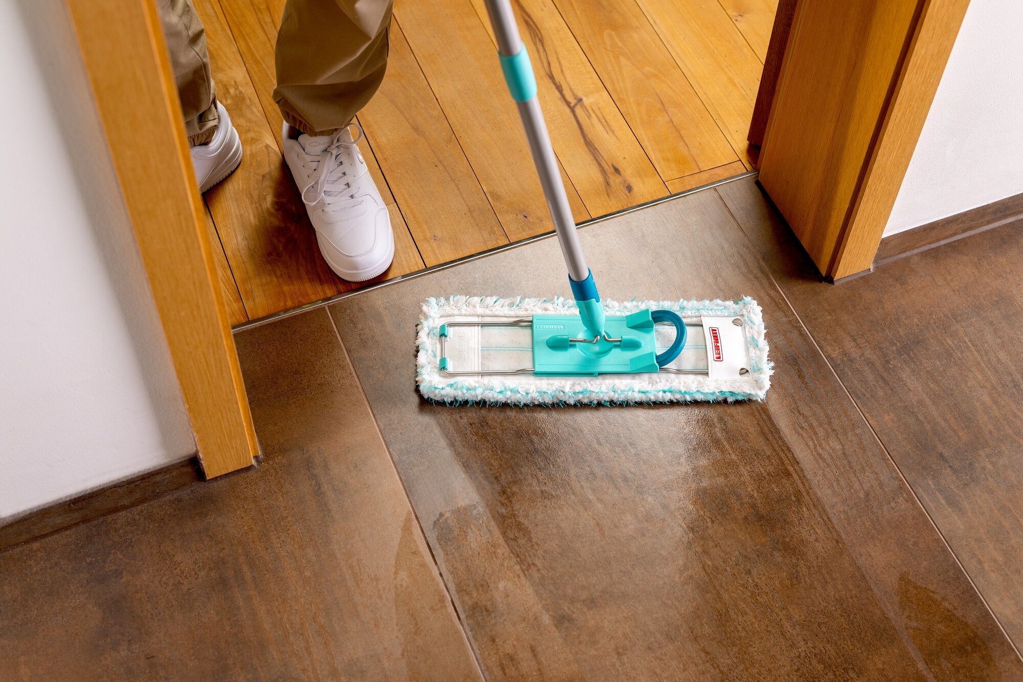Leifheit Profi XL Set avec presse à serpillière en cours d’utilisation lors du nettoyage humide d’un sol au niveau d’un seuil de porte. Le large balai de sol nettoie en profondeur les surfaces en bois et carrelées jusqu’au bord.