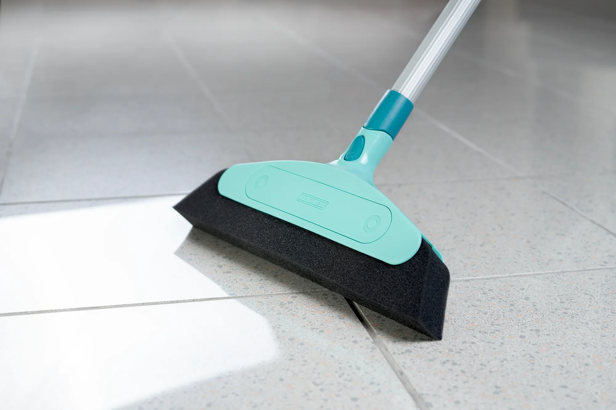 Balai en mousse avec système à clic sur un sol carrelé. Le balai a un manche turquoise et un bord en mousse noir, idéal pour les surfaces lisses.