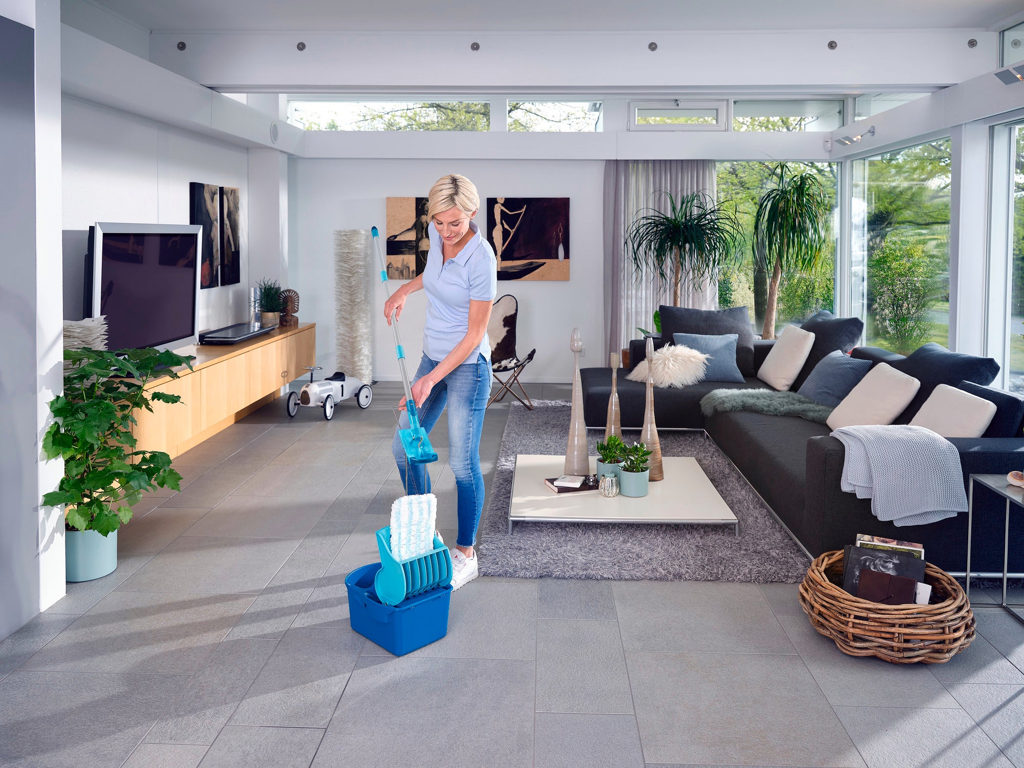 Une femme utilise la presse à essorger Profi Compact dans un salon moderne. La presse est posée sur le sol tandis que la femme verse de l'eau dans un récipient.