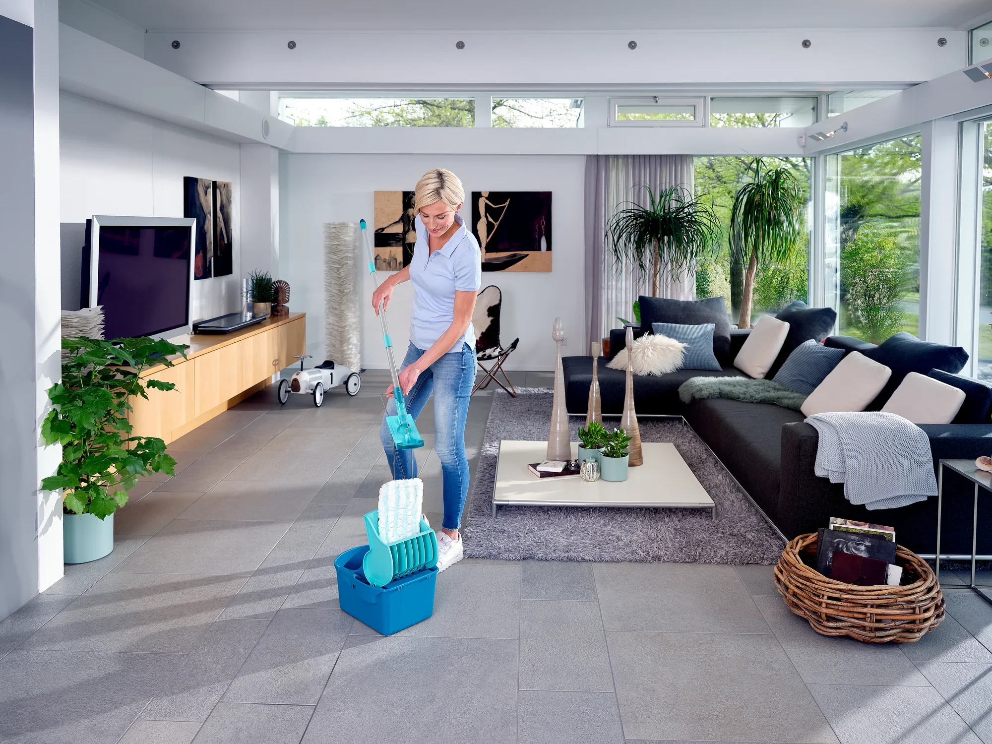 Een vrouw gebruikt de Profi Compact vloerwisser in een moderne woonkamer. De pers staat op de vloer terwijl de vrouw water uit een emmer giet.