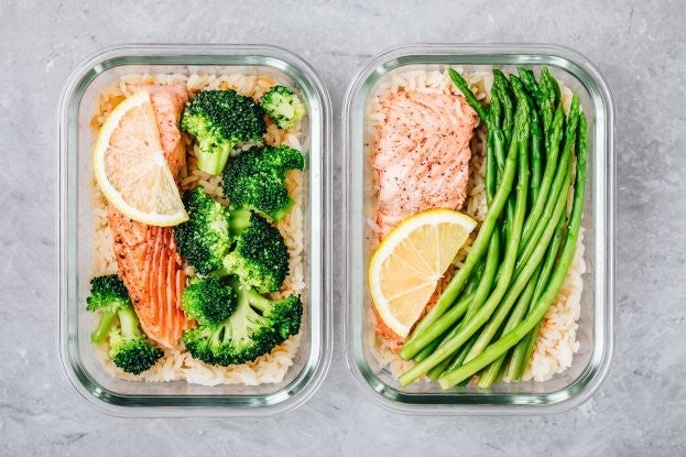 Zwei Glasbehaelter mit Reis, Lachs und Zitrone; links mit Brokkoli, rechts mit gruenem Spargel auf grauem Hintergrund.