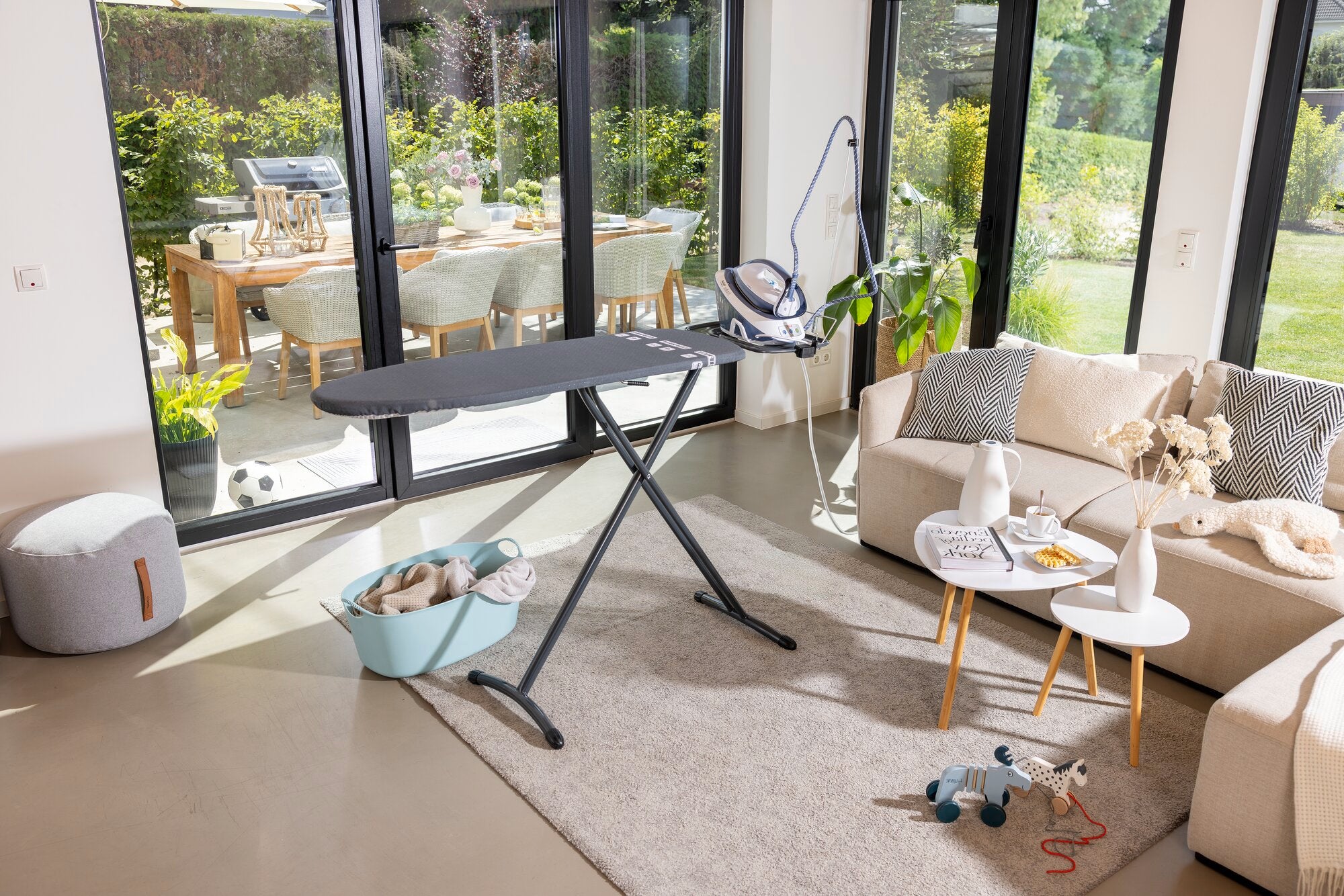 Bügelbrett Leifheit Air Board Express M Black in einem modernen Wohnzimmer. Das Brett steht auf einem Teppich, umgeben von Möbeln und Spielzeug, mit Blick auf einen sonnigen Garten.