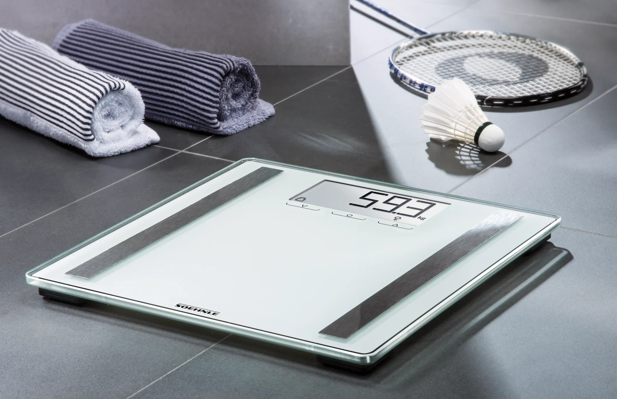 Cyfrowa waga łazienkowa, model Shape Sense Control 200, stoi na podłodze wyłożonej szarymi kafelkami. Obok leżą dwa zrolowane ręczniki i zestaw do badmintona.