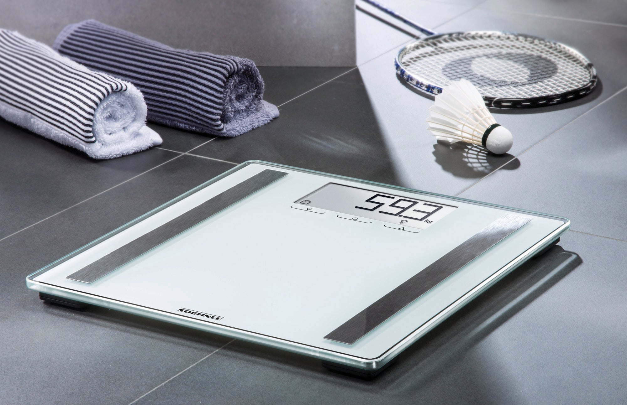 Eine Personenwaage digital, Modell Shape Sense Control 200, steht auf einem grauen Fliesenboden. Daneben liegen zwei gerollte Handtücher und ein Badminton-Set.