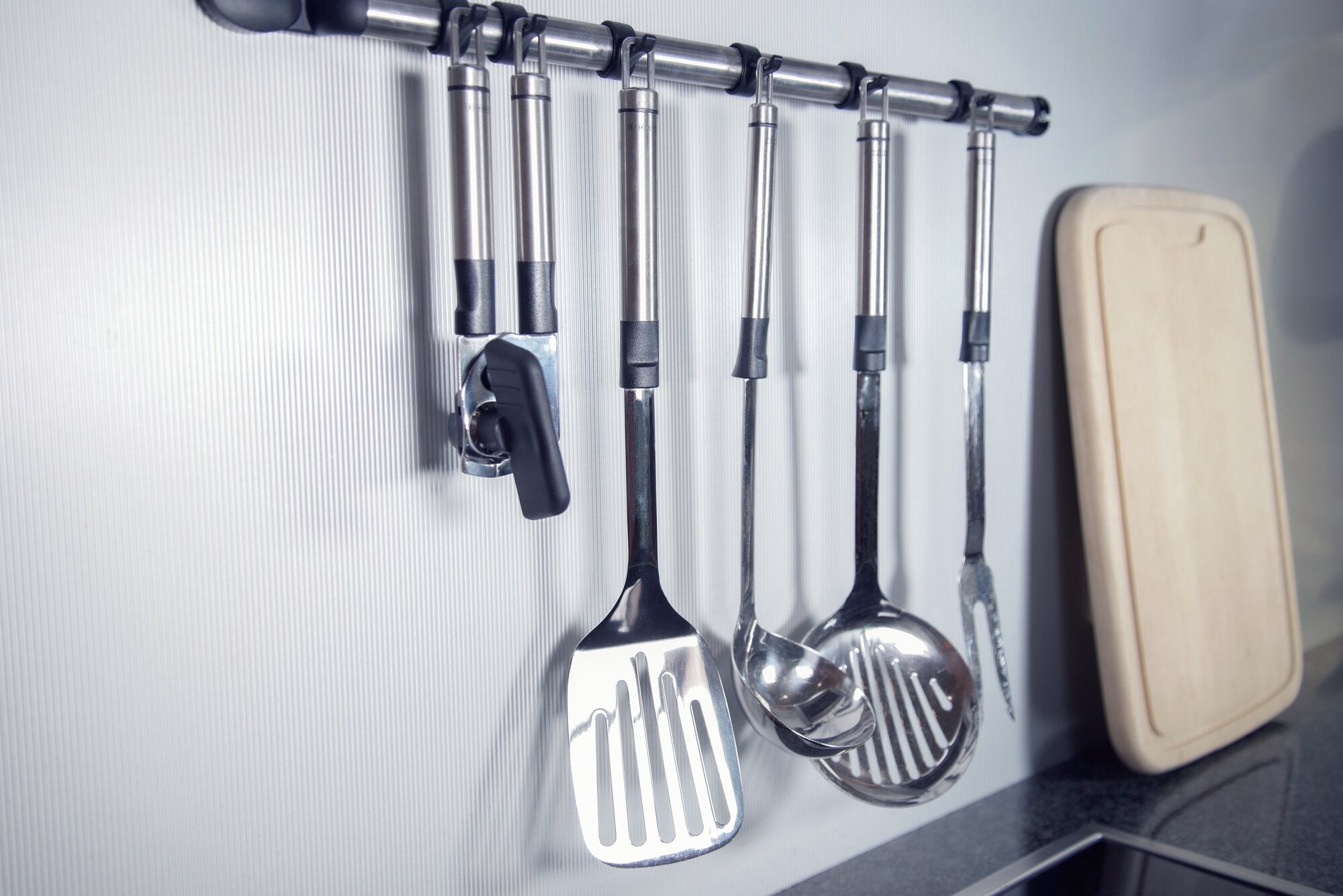 Un ouvre-boîte moderne est suspendu à une barre de cuisine à côté d'autres ustensiles de cuisine en acier inoxydable tels qu'une spatule et une louche. La scène montre un aménagement de cuisine élégant et organisé.