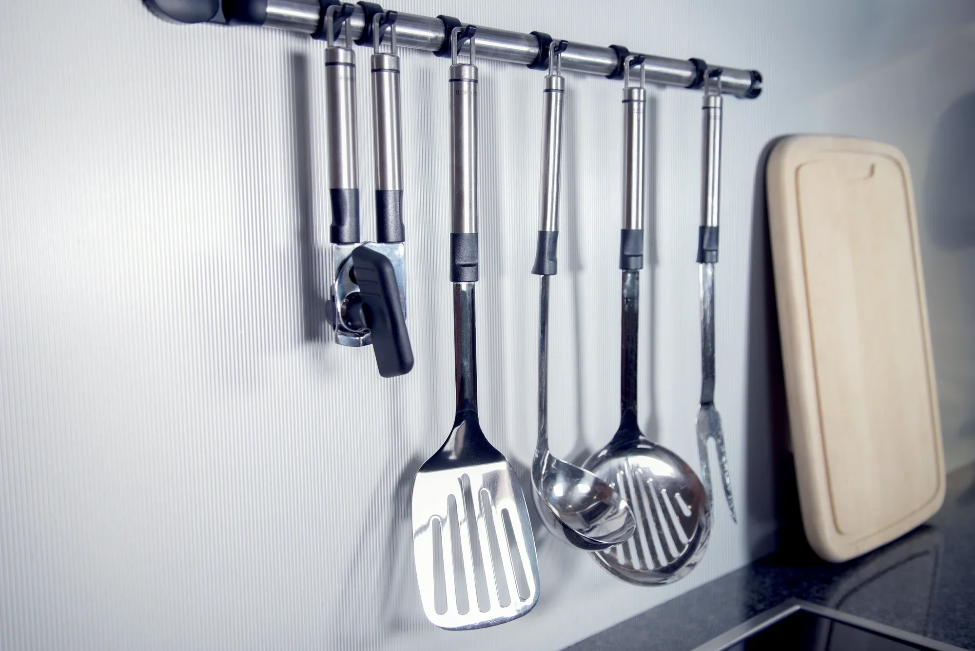 Nowoczesny otwieracz do puszek wisi na półce kuchennej obok innych przyborów kuchennych ze stali nierdzewnej, takich jak szpatułka i chochla do zupy. Scena przedstawia stylowe i uporządkowane wnętrze kuchni.