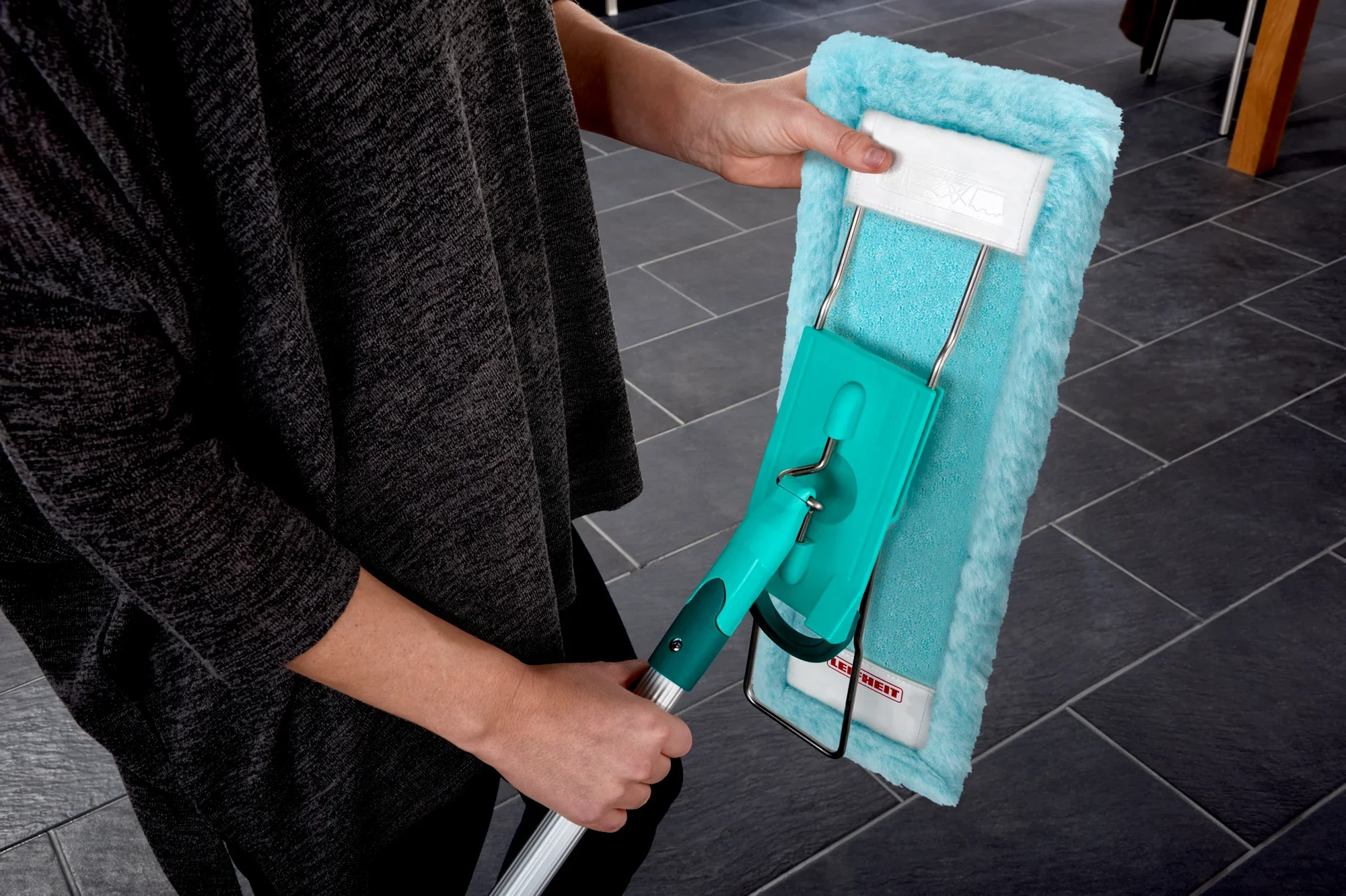 Une personne tient une housse de balai à franges modèle Profi XL. La housse est turquoise et s'utilise sur un sol carrelé foncé. Idéale pour un nettoyage en profondeur et une utilisation facile.