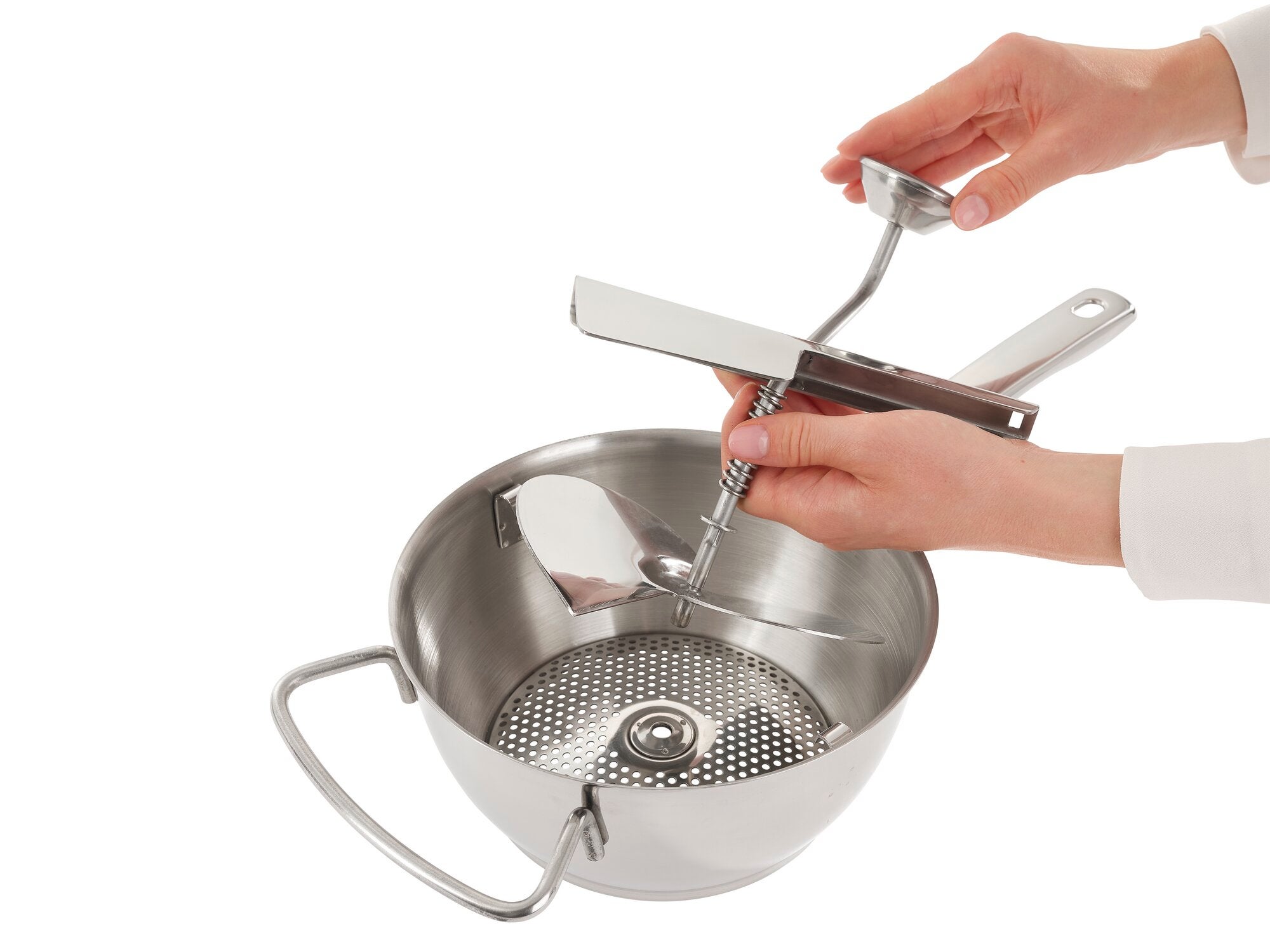 A person uses a stainless steel food mill to purée food. The mill has a sturdy handle and a crank that makes puréeing easy. Ideal for soups and sauces.