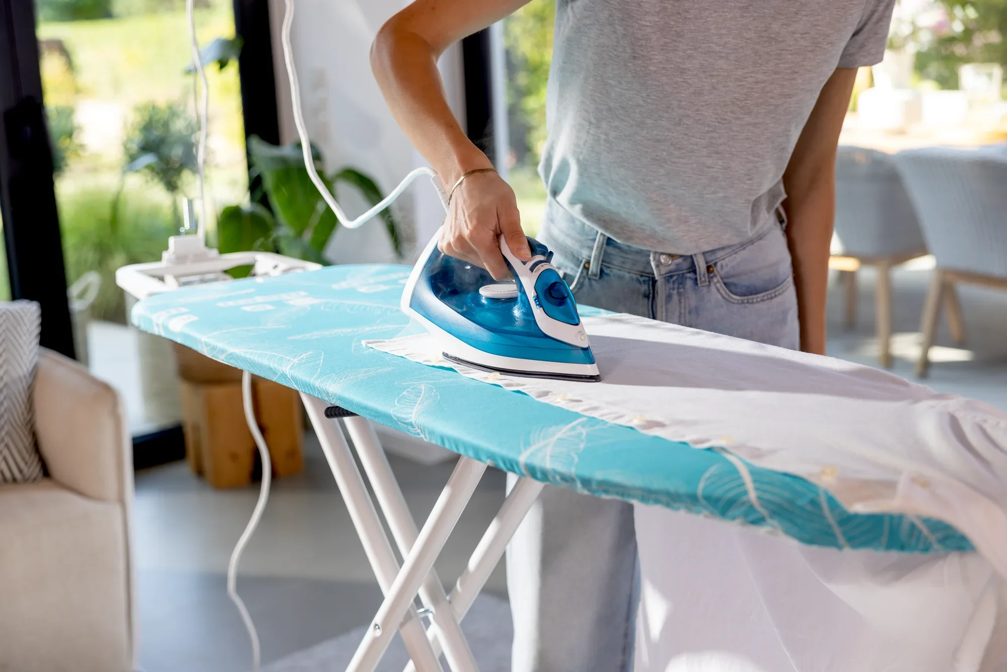 Eine Person bügelt ein Kleidungsstück auf dem Leifheit Air Board M Compact Plus Bügelbrett. Das Bügelbrett hat einen blauen Bezug und steht in einem hellen Raum.