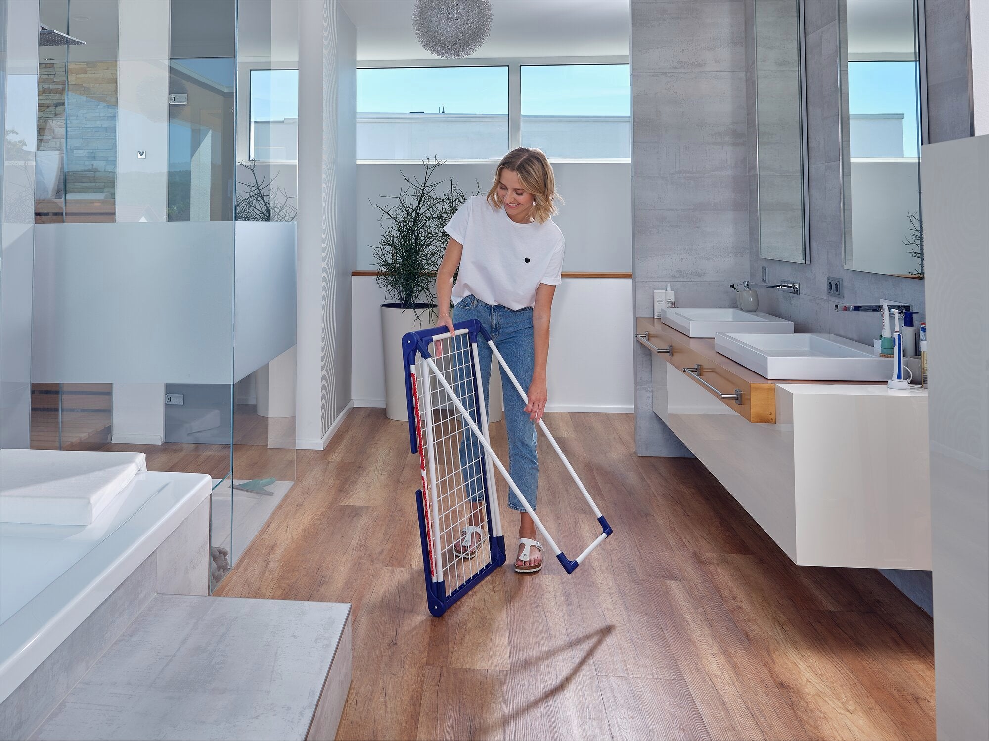 A woman stands in a modern bathroom holding the Pegasus 180 Solid Classic free-standing dryer. The dryer is folded up, showing its compact, space-saving design.
