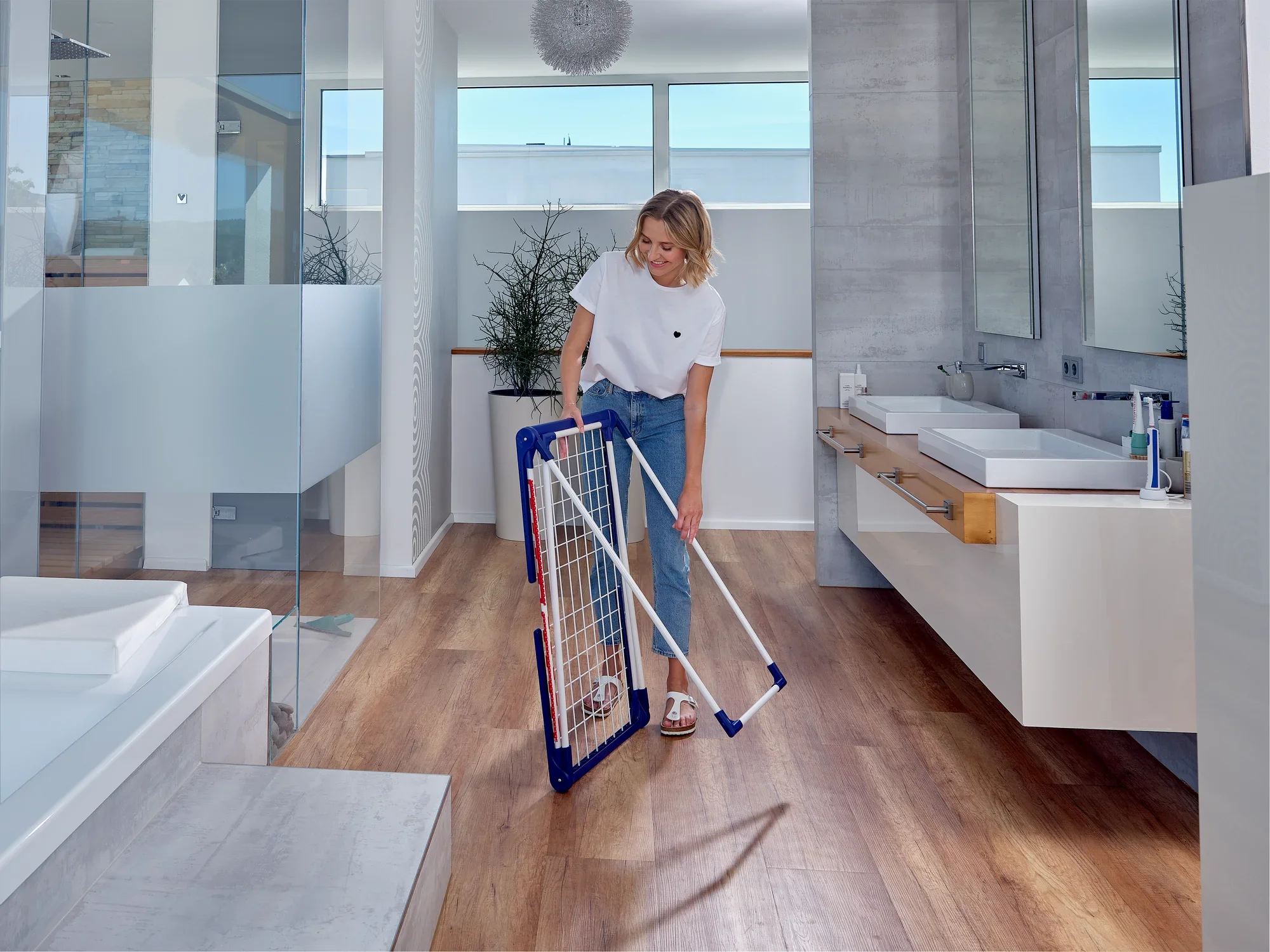 Kobieta stoi w nowoczesnej łazience i trzyma suszarkę bębnową Pegasus 180 Solid Classic. Suszarka jest złożona, dzięki czemu zajmuje niewiele miejsca.