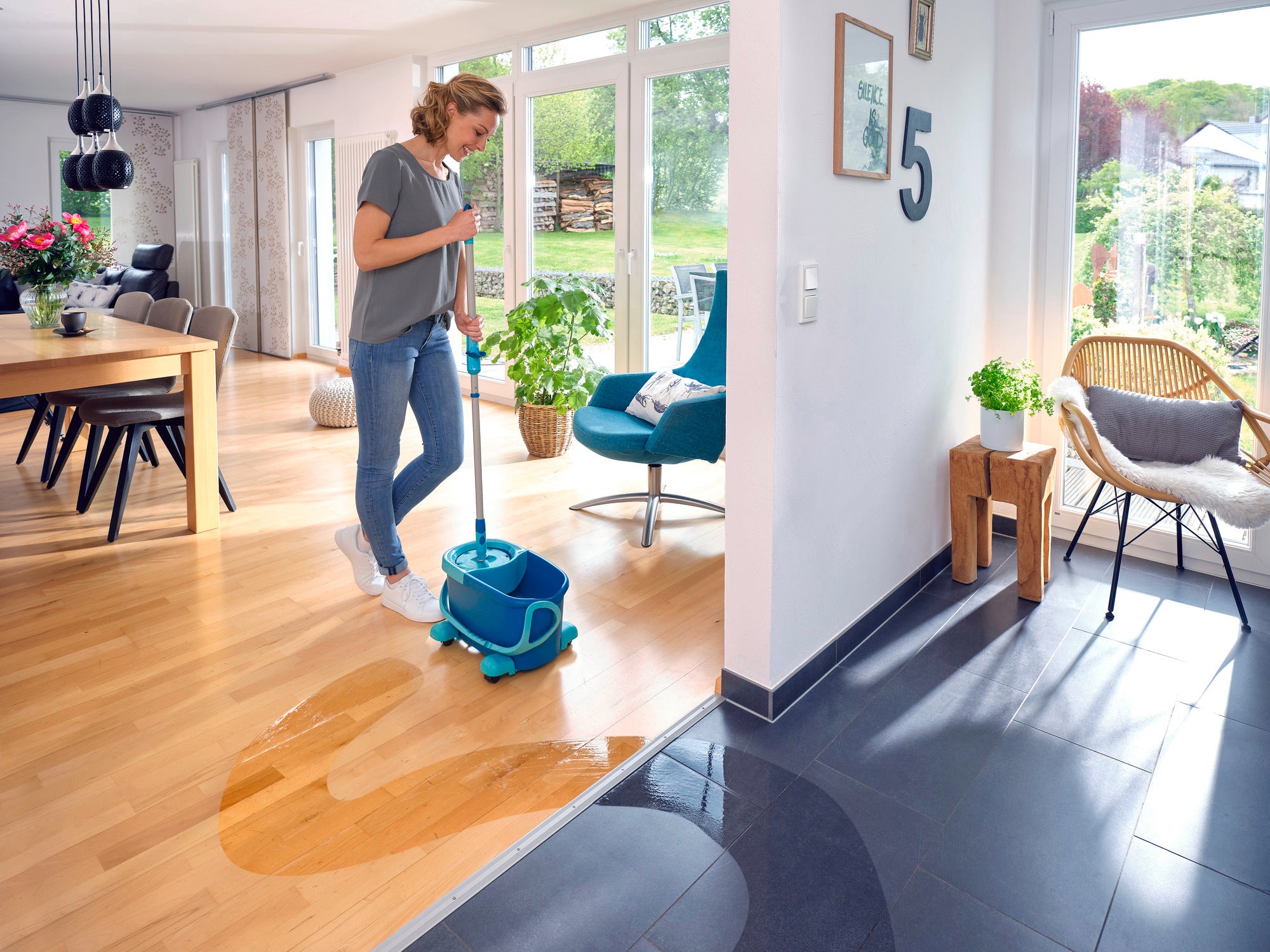 Une femme nettoie le sol en bois d'un salon moderne avec la serpillière CLEAN TWIST Disc Mop Ergo. La serpillière se trouve dans un seau bleu à roulettes, idéal pour un nettoyage efficace.