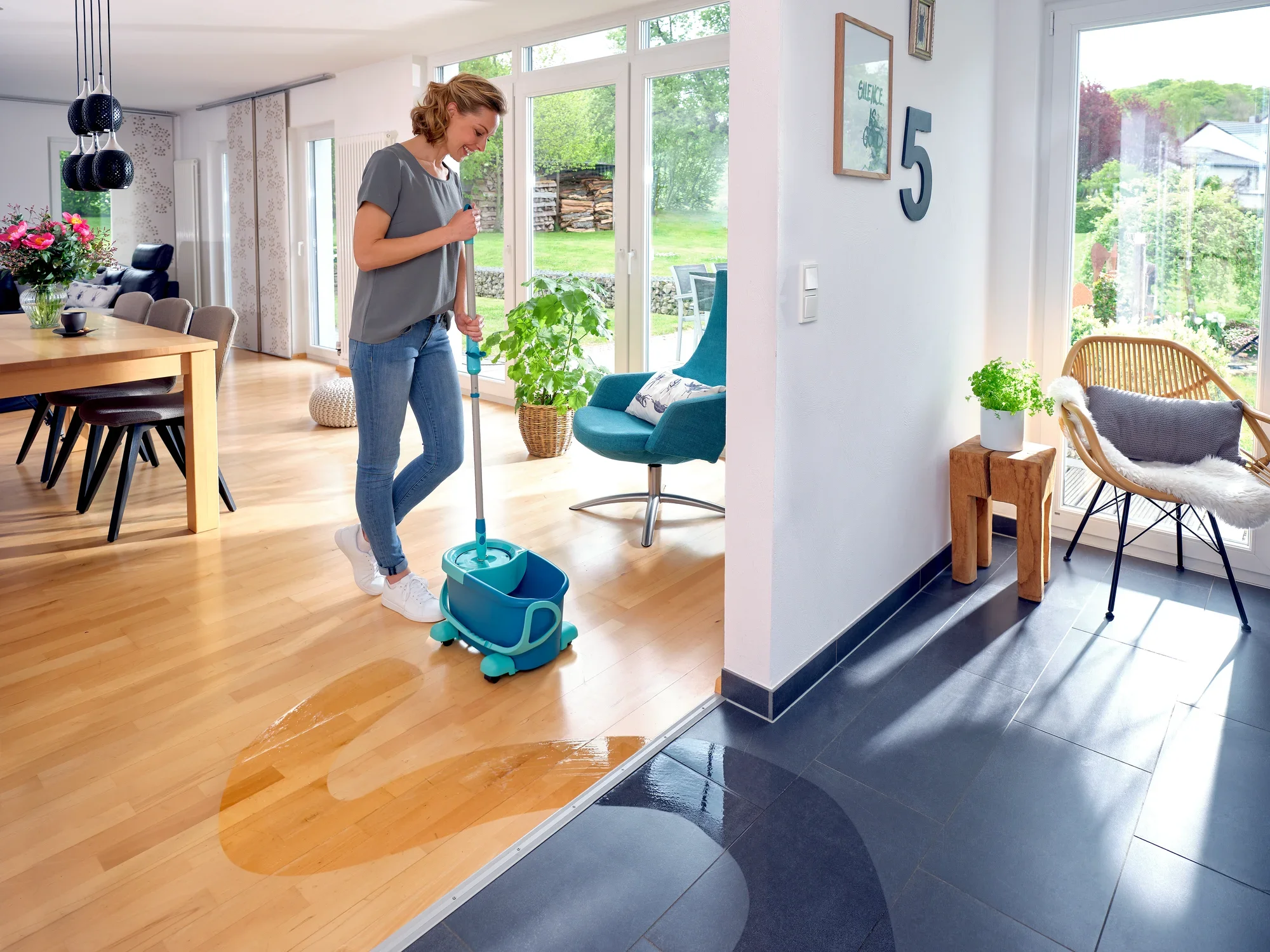 Een vrouw maakt de houten vloer van een moderne woonkamer schoon met de CLEAN TWIST Disc Mop Ergo. De mop wordt geleverd in een blauwe emmer met wieltjes, ideaal om efficiënt te dweilen.