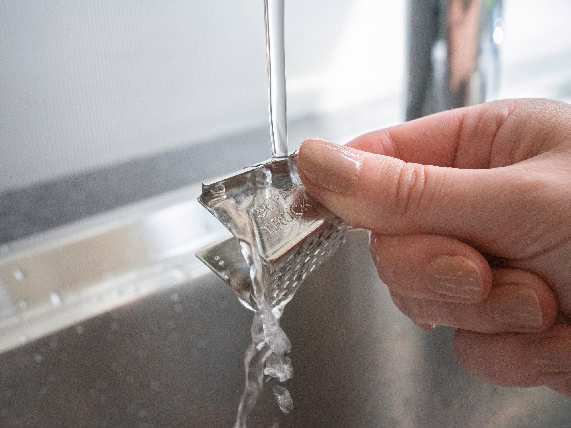 Eine Hand hält einen Edelstahl-Teebeutelhalter unter fließendem Wasser in einem Waschbecken. Der Halter hat eine strukturierte Oberfläche und wird gründlich gereinigt.