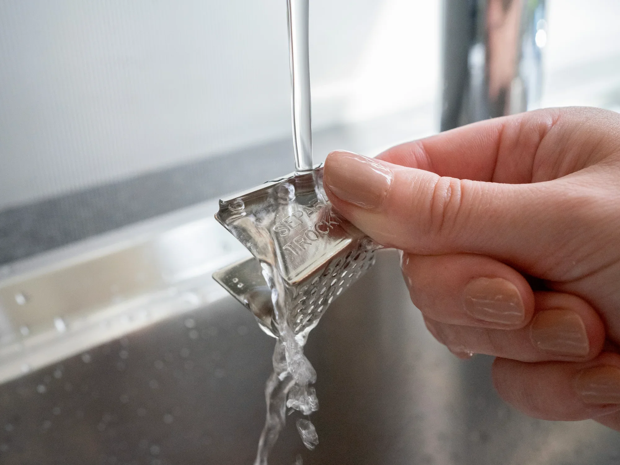 Uchwyt na torebki herbaty ze stali nierdzewnej trzymany jedną ręką pod bieżącą wodą w zlewie. Uchwyt ma teksturowaną powierzchnię i jest dokładnie czyszczony.