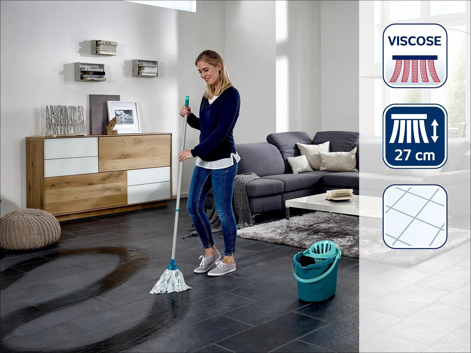 Una donna pulisce il pavimento di un moderno soggiorno con un lavapavimenti Leifheit. La stanza è arredata con stile, con un divano grigio e mobili in legno. Accanto a lei c'è un secchio.