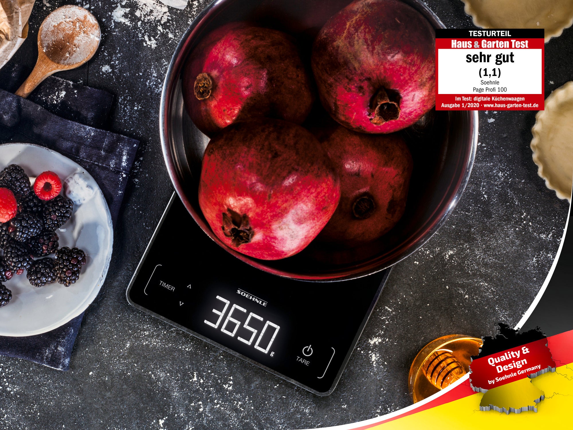 A digital kitchen scale shows 3650 grams while weighing a bowl of pomegranates. Next to it are berries and honey. The scale has received an award for quality and design.