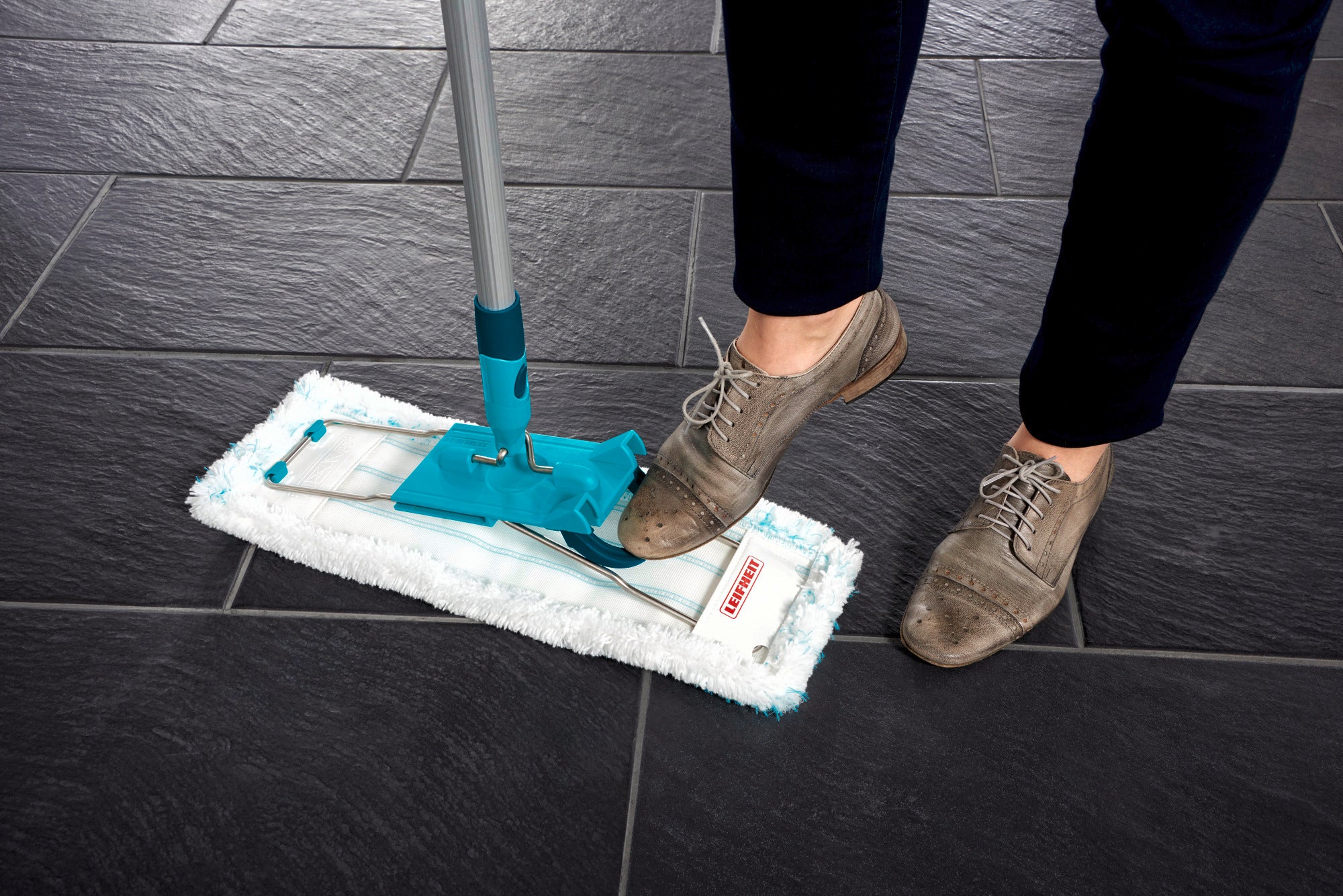 Eine Person benutzt den Bodenwischer Profi XL auf dunklen Fliesen. Der Wischer hat ein blaues Gelenk und ein weißes Mikrofasertuch, das für gründliche Reinigung sorgt.