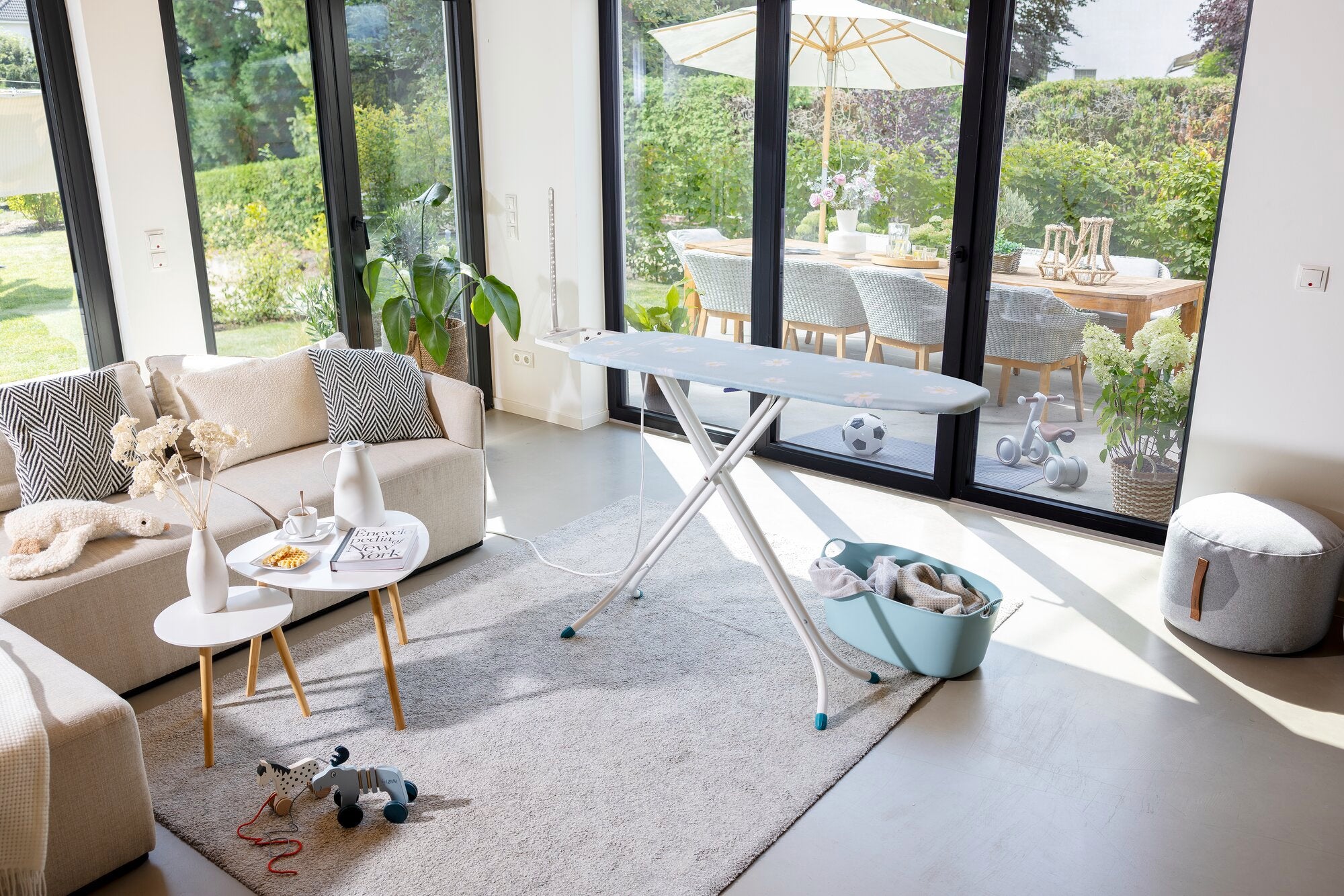 Bügelbrett Leifheit Classic M Compact Plus in einem modernen Wohnzimmer mit großen Fenstern. Das Brett steht auf einem hellen Teppich, umgeben von stilvollen Möbeln und Dekorationen.