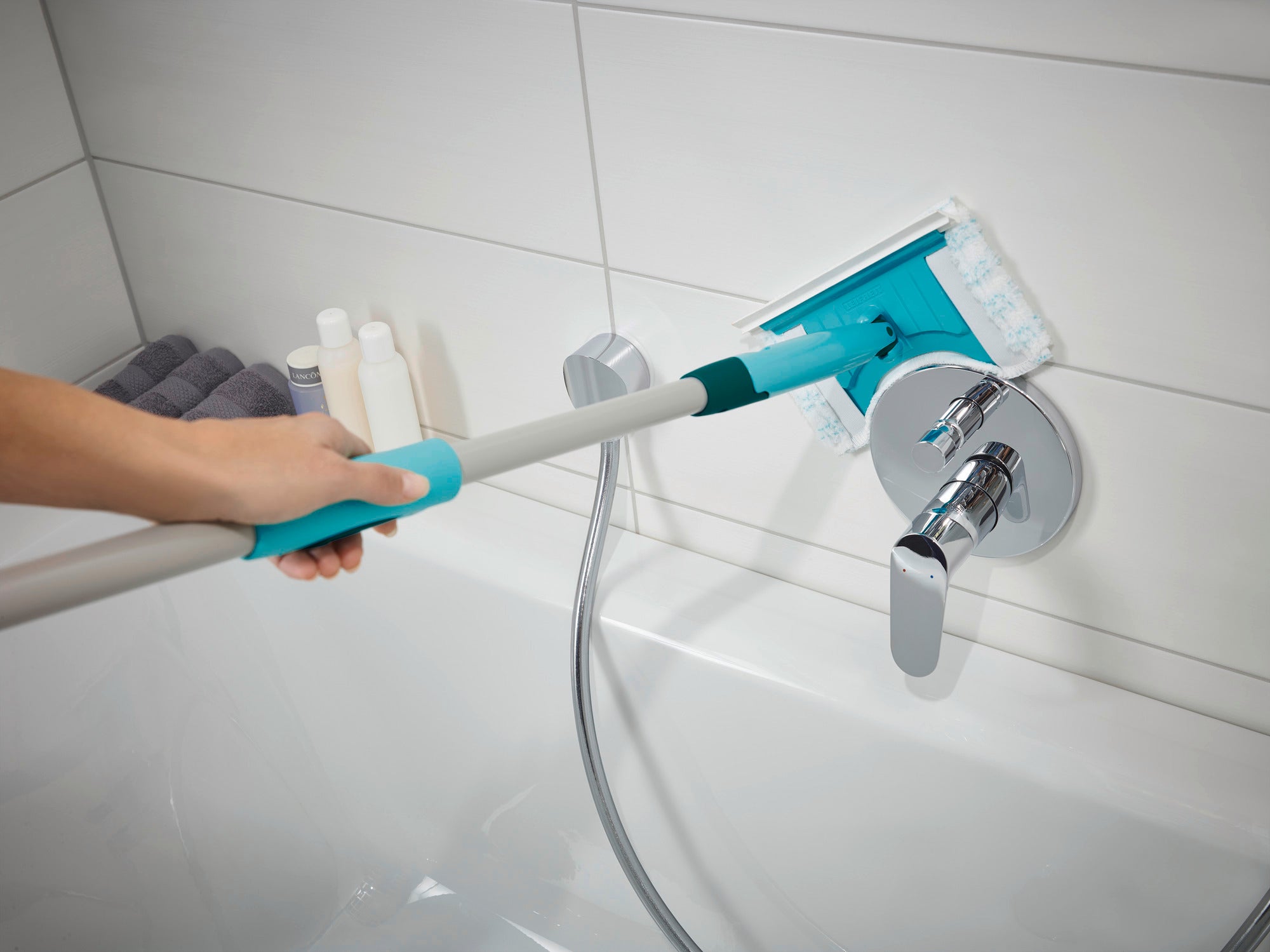 Fliesen- und Wannenwischer Flexipad mit Teleskopstiel wird in einem Badezimmer verwendet. Eine Hand hält den Wischer, während er über eine weiße Fliese gleitet. Im Hintergrund sind Pflegeprodukte sichtbar.