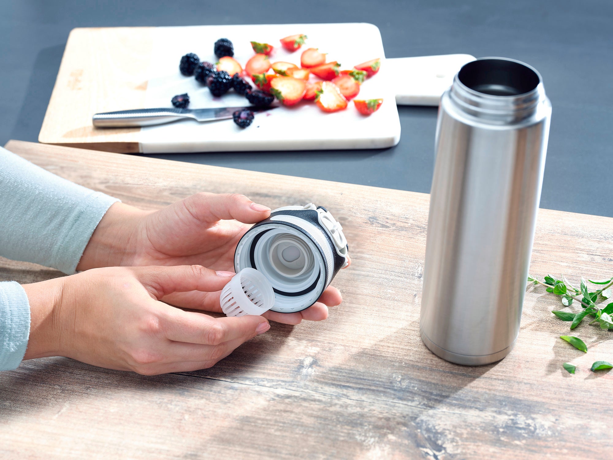 Geïsoleerde mok Flip 600ml in zilver op een tafel. Eén persoon houdt het deksel van de mok vast terwijl op de achtergrond vers fruit op een snijplank ligt.