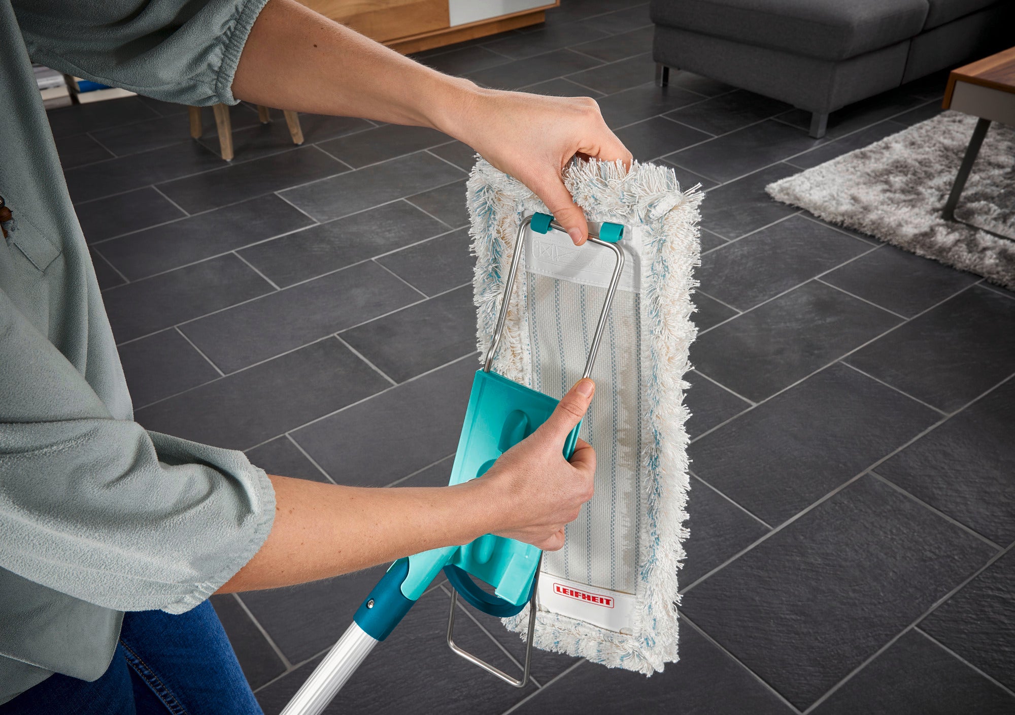 Eine Person benutzt den Bodenwischer Profi XL auf einem dunklen Fliesenboden. Der Wischer hat einen grünen Griff und ein weißes, saugfähiges Wischtuch, das für gründliche Reinigung sorgt.