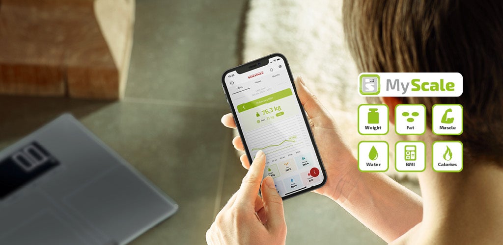 Ein Mensch haelt ein Smartphone mit der MyScale-App, die Gewichtsdaten und Diagramme anzeigt, im Hintergrund liegt eine Waage.