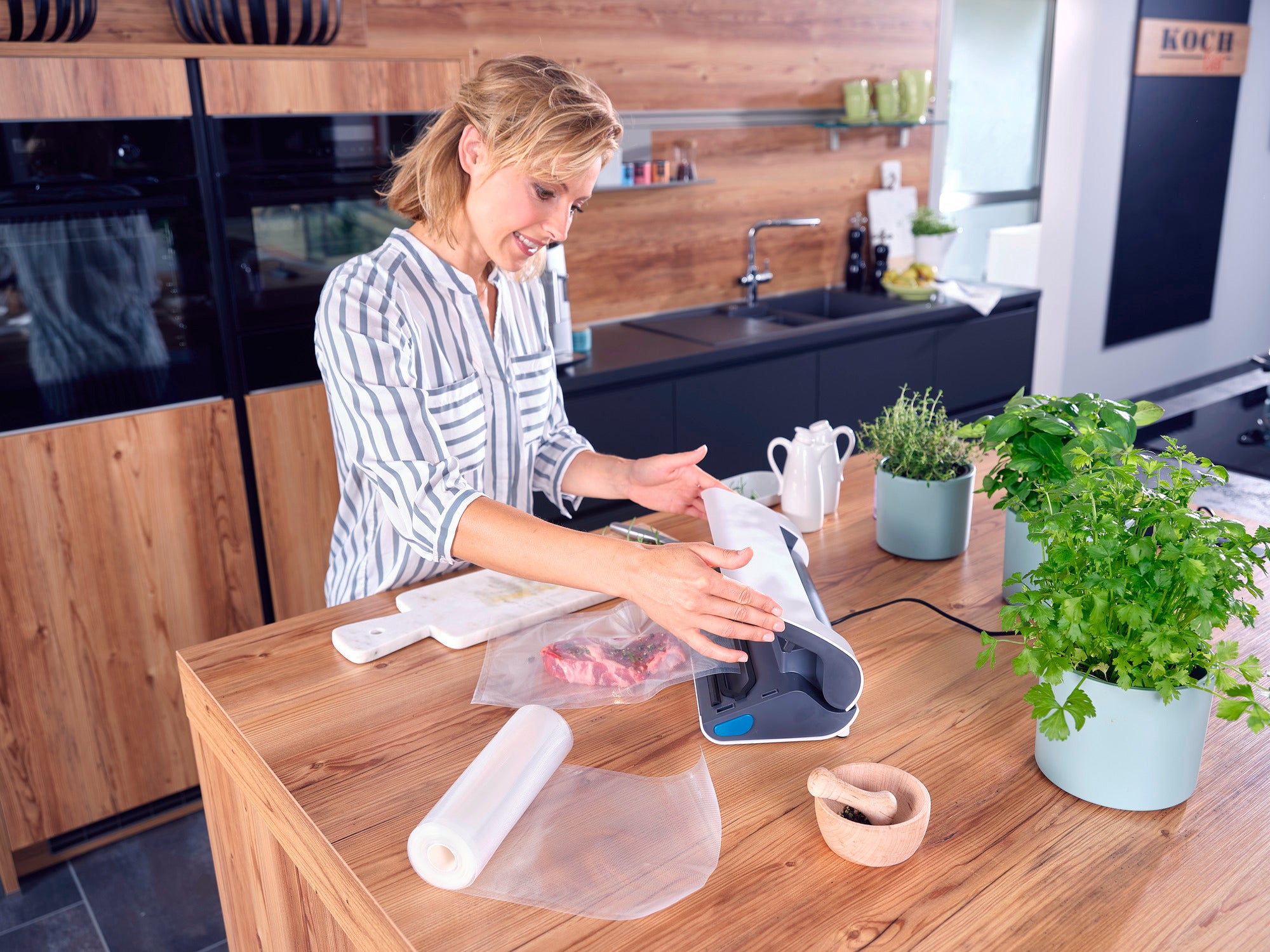 Eine Frau benutzt das Vakuumiergerät Vacu Power 300 in einer modernen Küche. Sie vakuumiert Lebensmittel, während frische Kräuter und Küchenutensilien auf der Arbeitsplatte stehen.