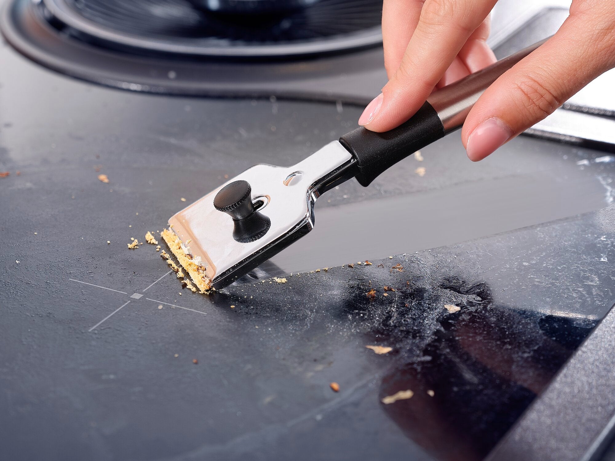 Une main utilise un grattoir de plaque de cuisson pour enlever les restes de nourriture d'une plaque de cuisson lisse. Le grattoir a un manche noir et une lame en métal. Idéal pour nettoyer les plaques de cuisson.