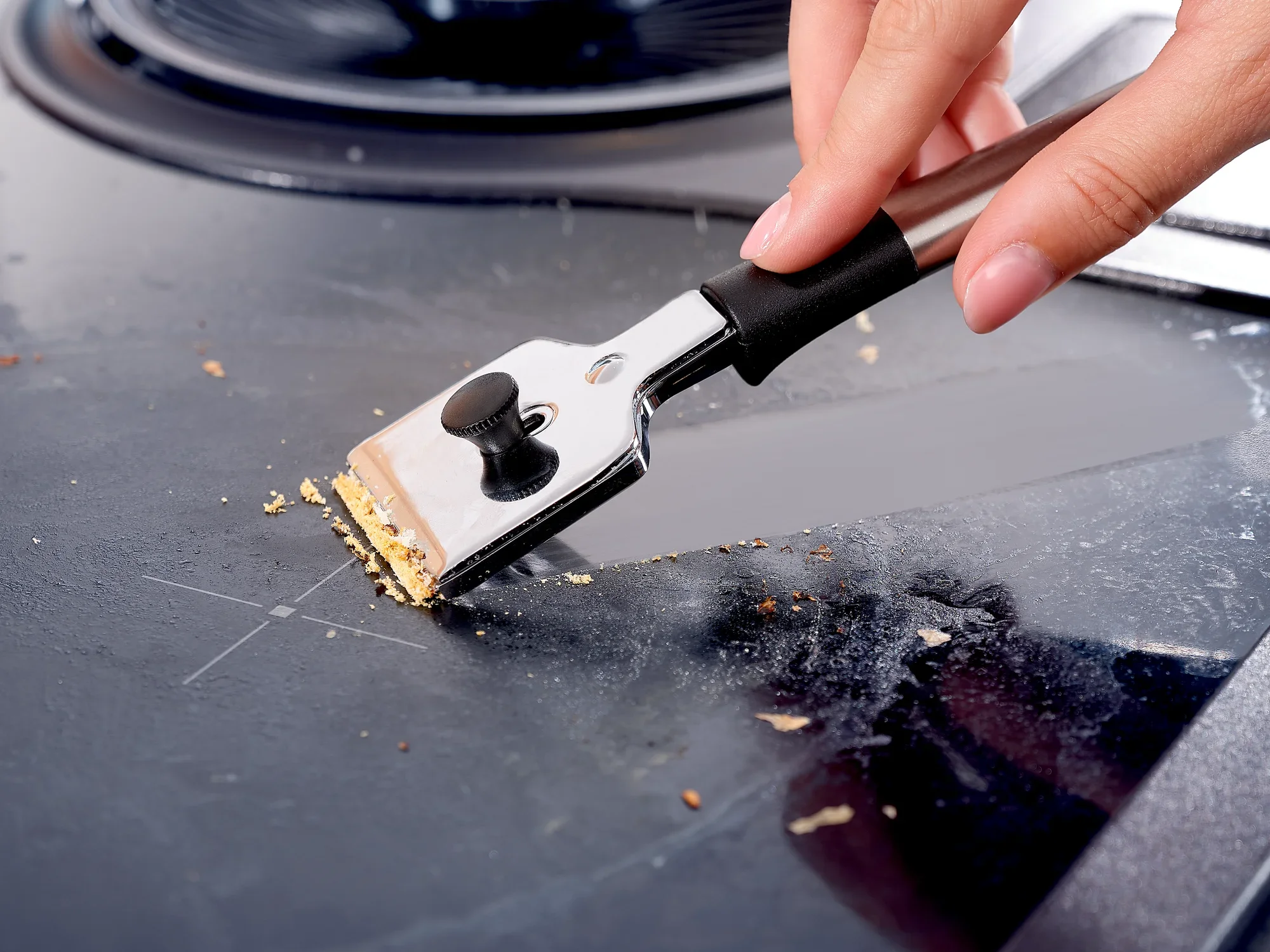 Jedna ręka używa skrobaka do płyty kuchennej, aby usunąć resztki jedzenia z gładkiej płyty. Skrobak ma czarny uchwyt i metalowe ostrze. Idealny do czyszczenia płyt grzewczych.