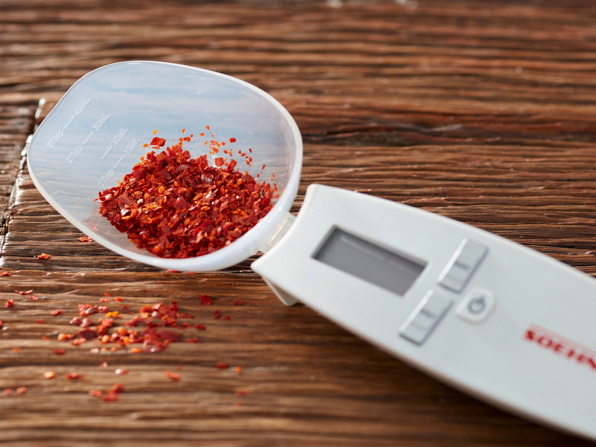 Balance de cuisine numérique Cooking Star avec écran, posée sur une table en bois. La balance est équipée d'un plateau amovible rempli de flocons d'épices rouges. Idéale pour peser avec précision.