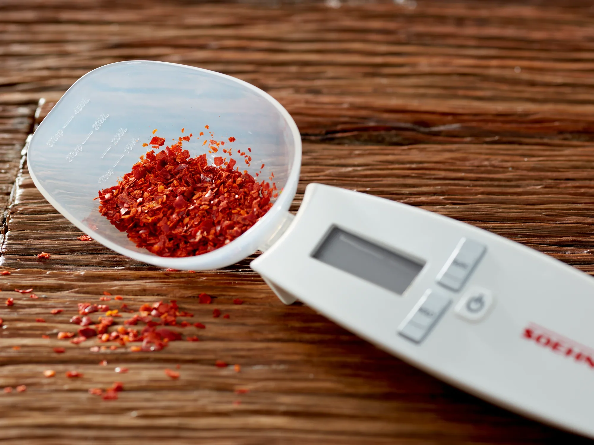 Cyfrowa waga kuchenna Cooking Star z wyświetlaczem, leżąca na drewnianym stole. Waga ma wyjmowaną miskę wypełnioną czerwonymi płatkami przypraw. Idealna do precyzyjnego ważenia.