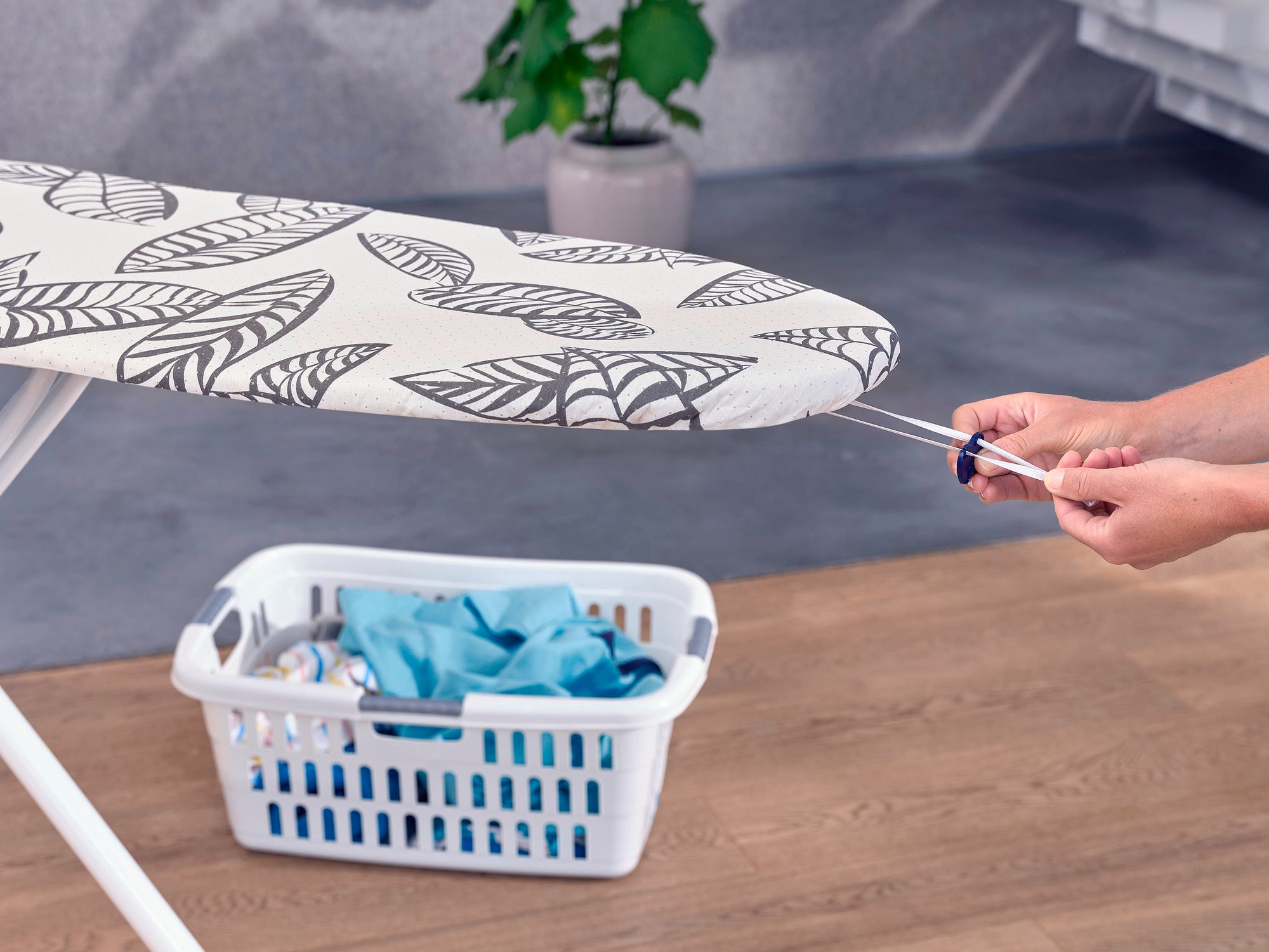 Housse pour table à repasser Perfect Steam : gros plan d'une table à repasser avec une housse à motifs floraux. Une personne tire sur une bande élastique pour fixer la housse. Un panier à linge se trouve à l'arrière-plan.