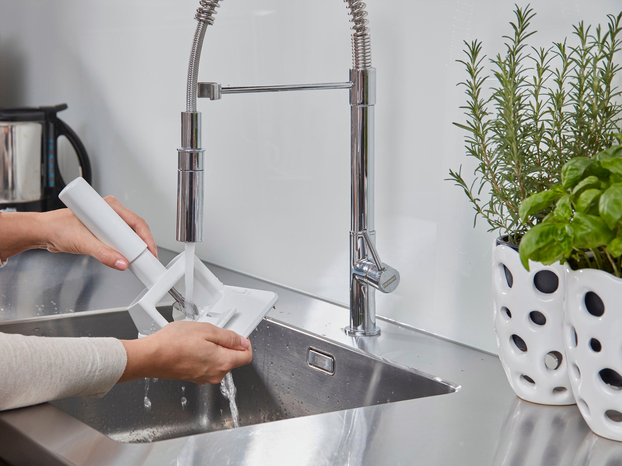 Een persoon maakt een pruimenpitter schoon onder stromend water in een moderne keuken. De ontpitter is wit en wordt afgespoeld onder een glimmende kraan.