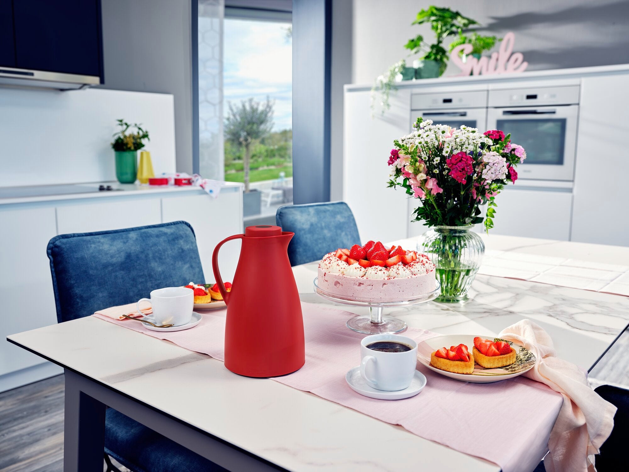 Isolierkanne Harmonic in einem modernen Esszimmer auf einem Tisch mit einer Torte, Blumenstrauß und zwei Tassen Kaffee. Die Kanne ist rot und steht im Mittelpunkt der Szene.