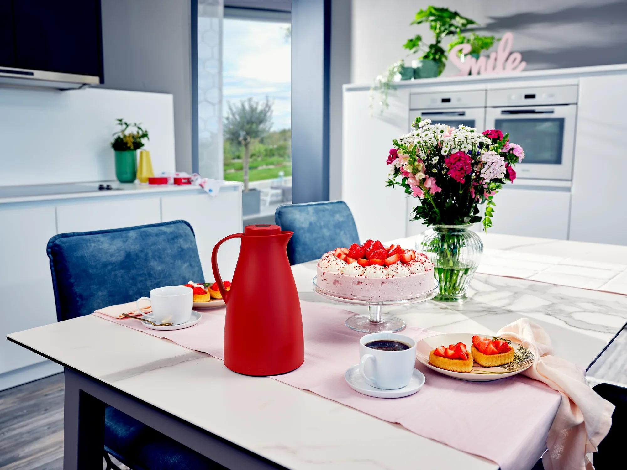 Harmonic isoleerkan in een moderne eetkamer op een tafel met een taart, een boeket bloemen en twee kopjes koffie. De kan is rood en vormt het middelpunt van het tafereel.