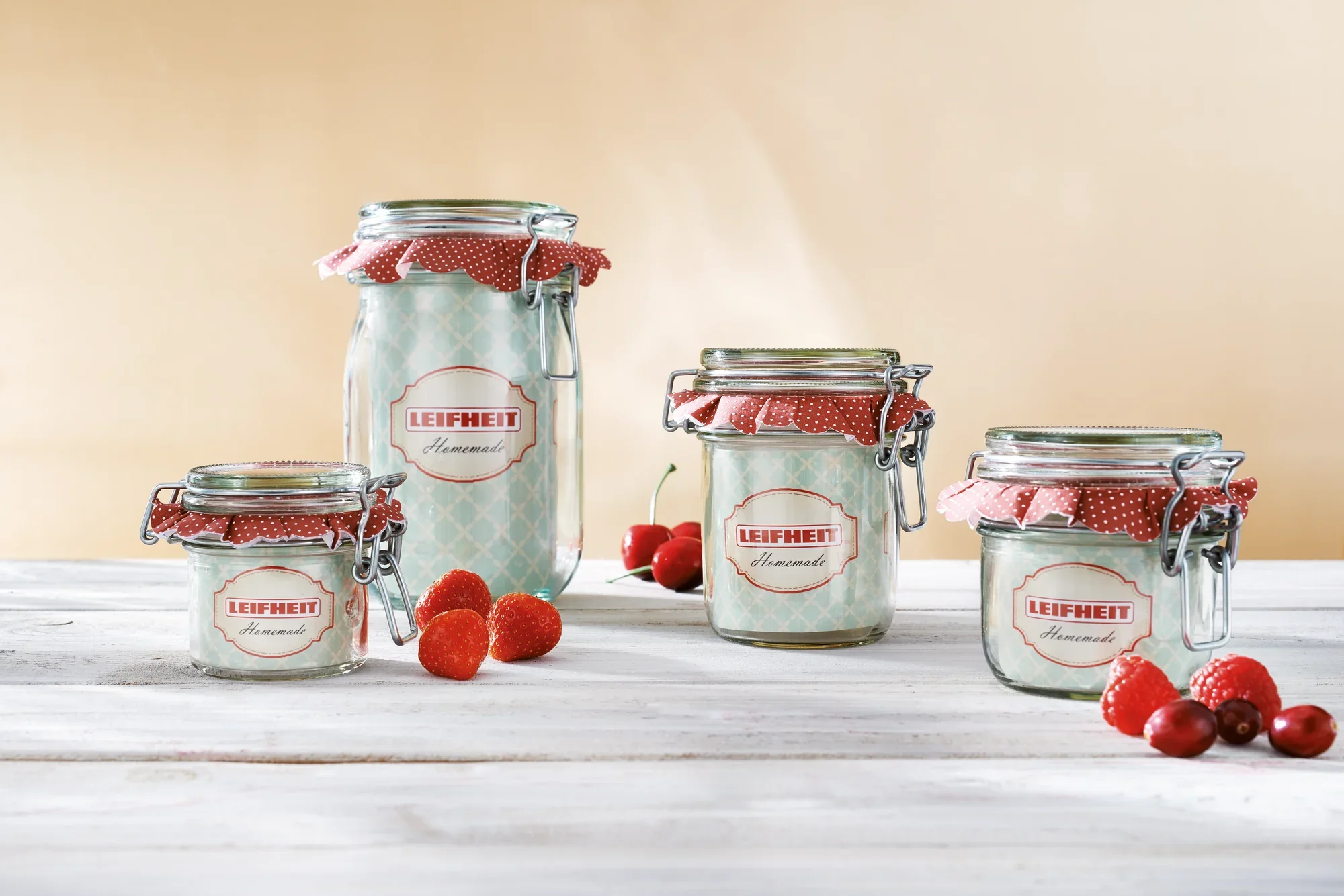 Cztery druciane słoiki Leifheit w różnych rozmiarach stoją na drewnianym stole. Są one przykryte pokrywkami z czerwonego materiału. Na pierwszym planie znajdują się świeże truskawki i maliny.