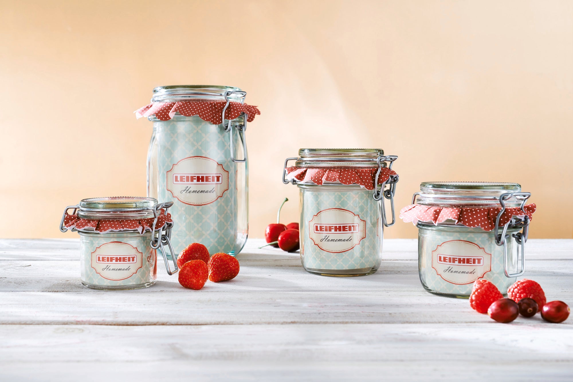 Cuatro tarros con cierre de alambre de Leifheit en diferentes tamaños sobre una mesa de madera. Están cubiertos con tapas de tela roja. En primer plano hay fresas y frambuesas frescas.
