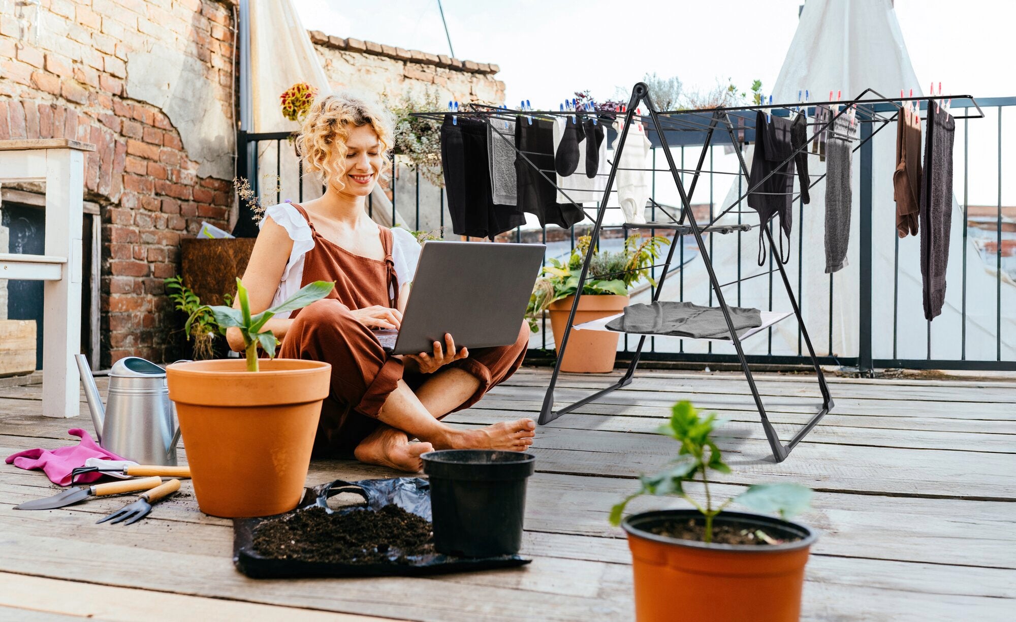 A woman sits on a terrace working on a laptop. Next to her is the Classic 250 Flex Black free-standing dryer with clothes hanging on it to dry. Plants and garden tools are also visible.