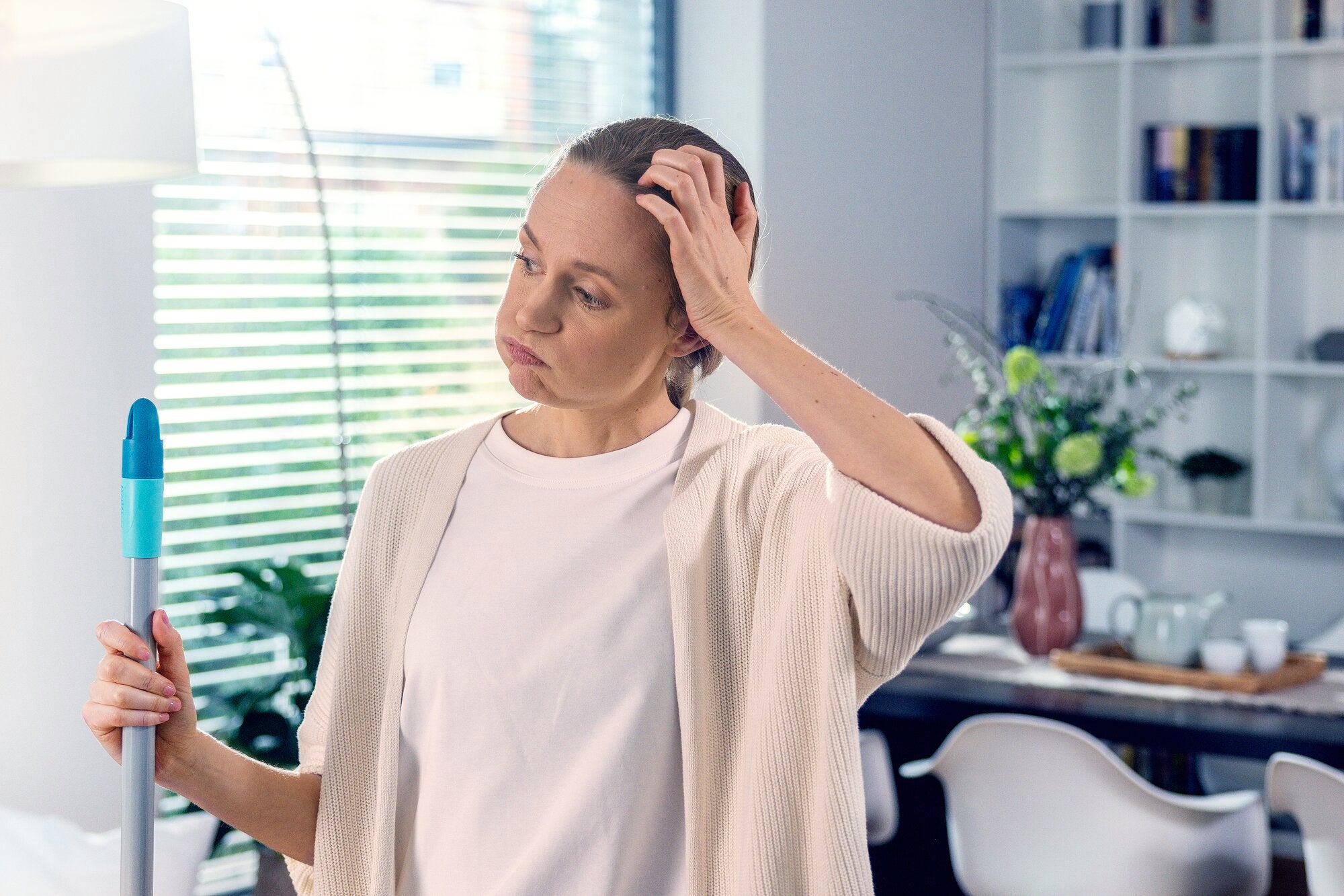 Eine Frau steht nachdenklich in einem modernen Wohnzimmer und hält einen POWER CLEAN Wischmopp. Im Hintergrund sind ein Esstisch und ein Bücherregal zu sehen.