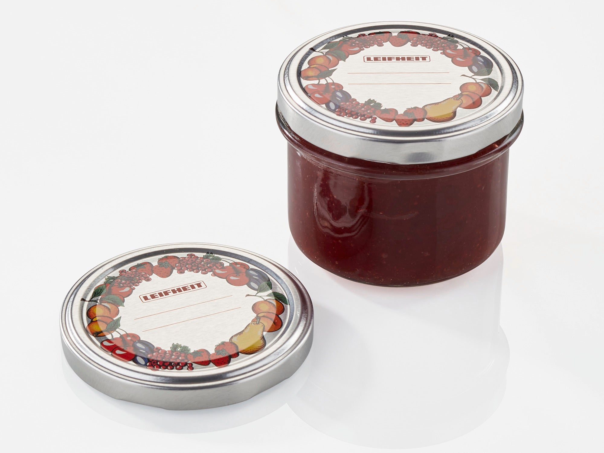 A jar with red contents and a removable replacement lid measuring 82 mm in diameter. The lid is decorated with a colourful fruit motif and lies next to the jar.
