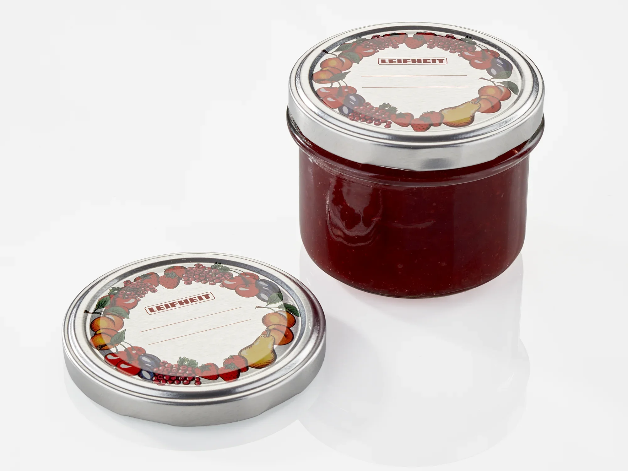 Słoik z czerwoną zawartością i zdejmowaną pokrywką o średnicy 82 mm. Pokrywka jest ozdobiona kolorowym motywem owoców i leży obok słoika.