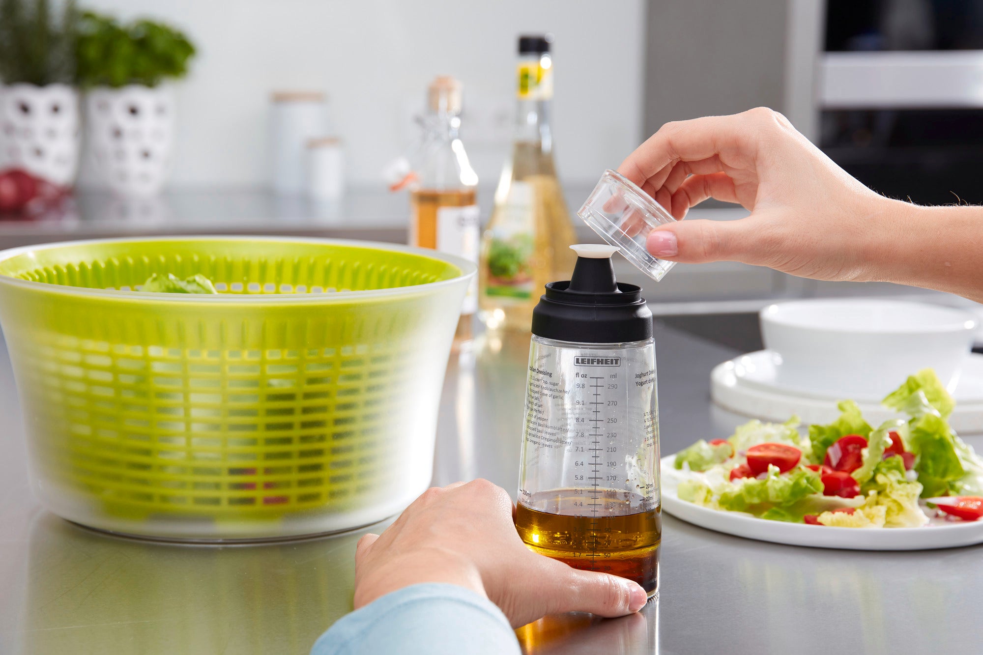 Ein Salat-Dressing-Shaker wird in einer modernen Küche verwendet. Eine Hand hält den Shaker, während eine andere Hand eine kleine Menge Dressing hinzufügt. Im Hintergrund ist eine Salatschleuder zu sehen.