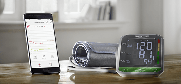 Ein Smartphone zeigt Blutdruckdaten, daneben ein elektronisches Blutdruckmessgeraet mit Manschette auf einem Holztisch.