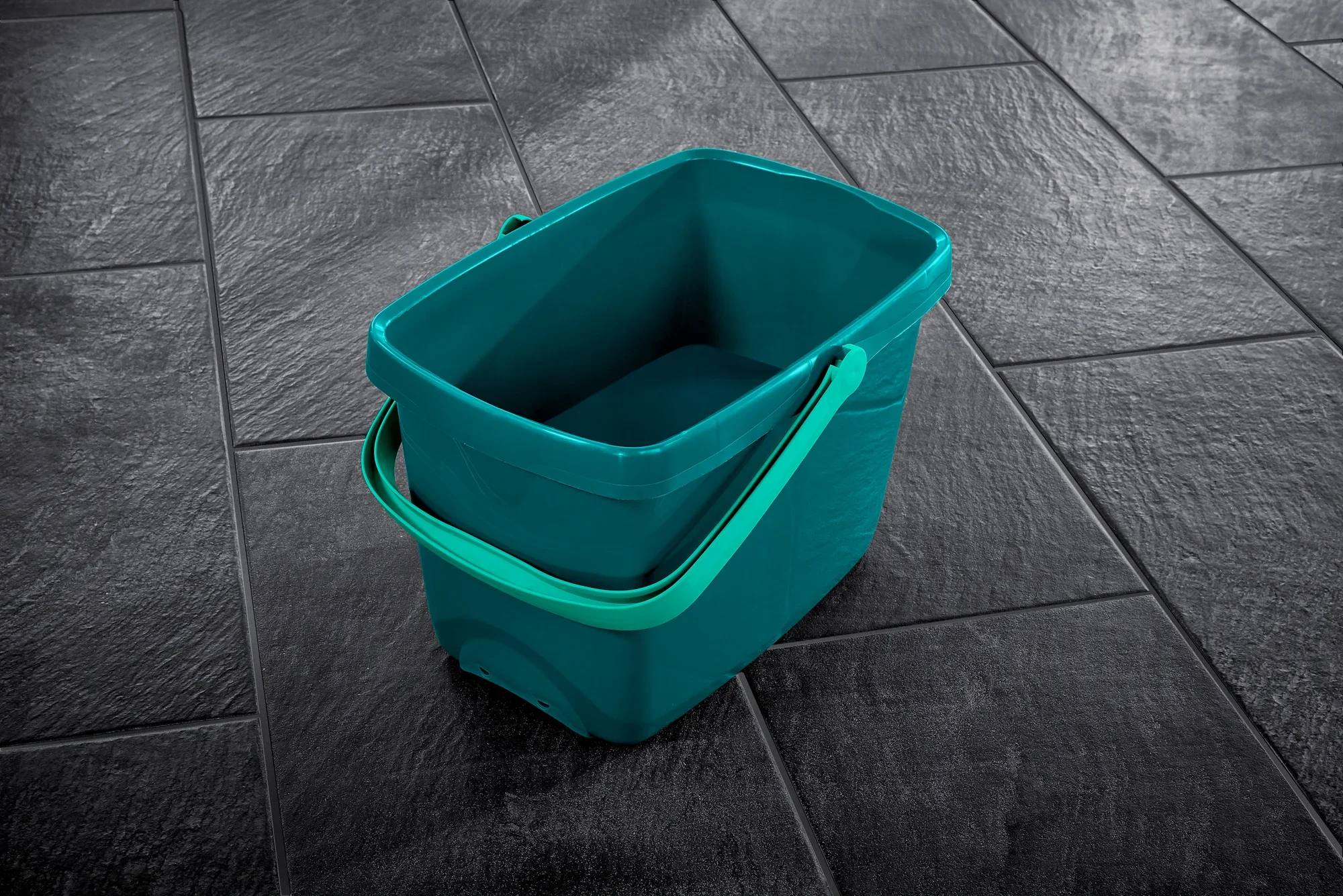 Un cubo azul con asa verde sobre un suelo de baldosas oscuras. El cubo es rectangular y ofrece espacio suficiente para las tareas de limpieza. Ideal para usar con el sistema Profi XL.