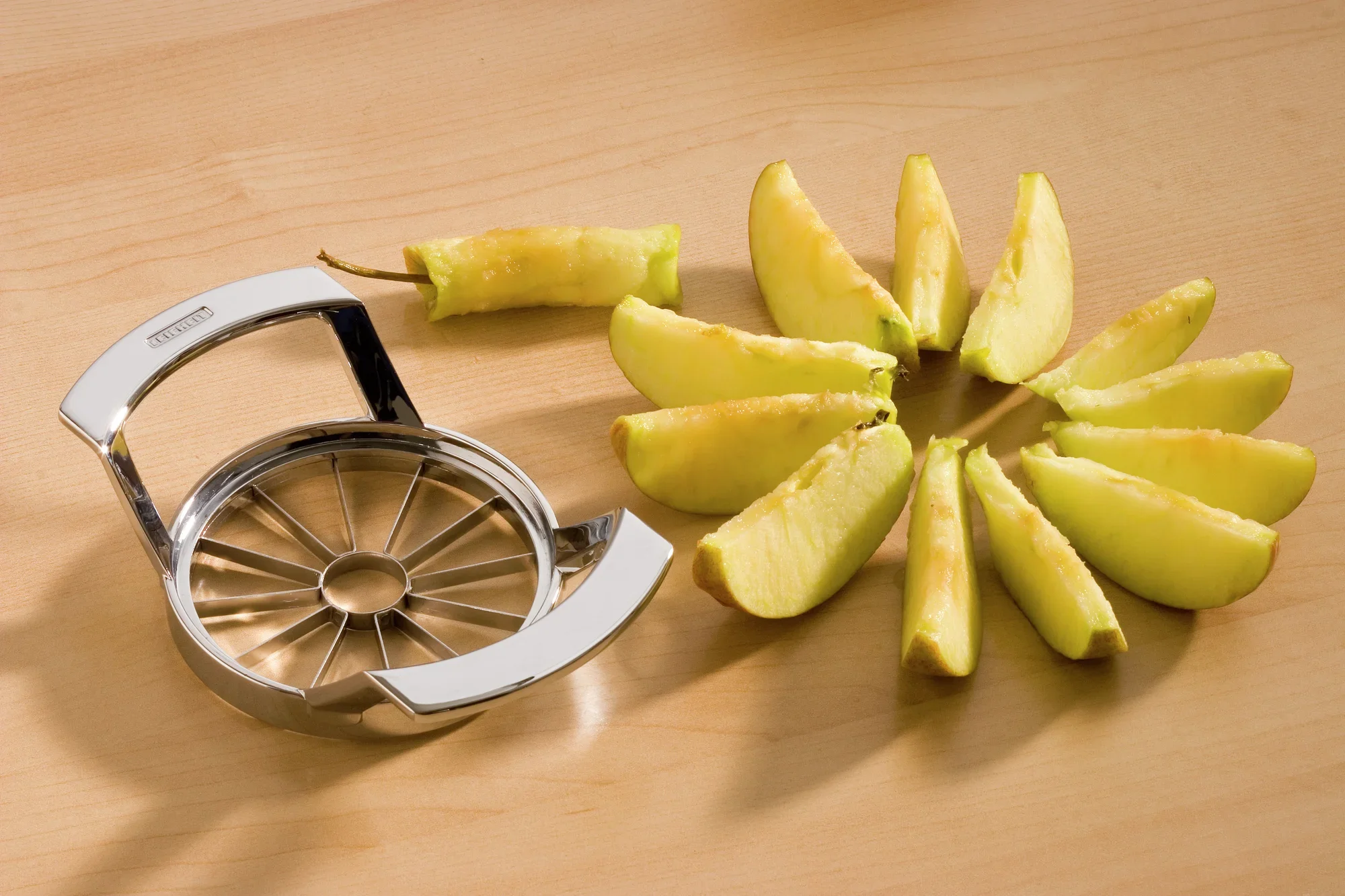 Krajalnica do jabłek ze stali nierdzewnej leży na drewnianym stole. Obok widać równo pokrojone kawałki jabłka, co demonstruje zastosowanie krajalnicy.