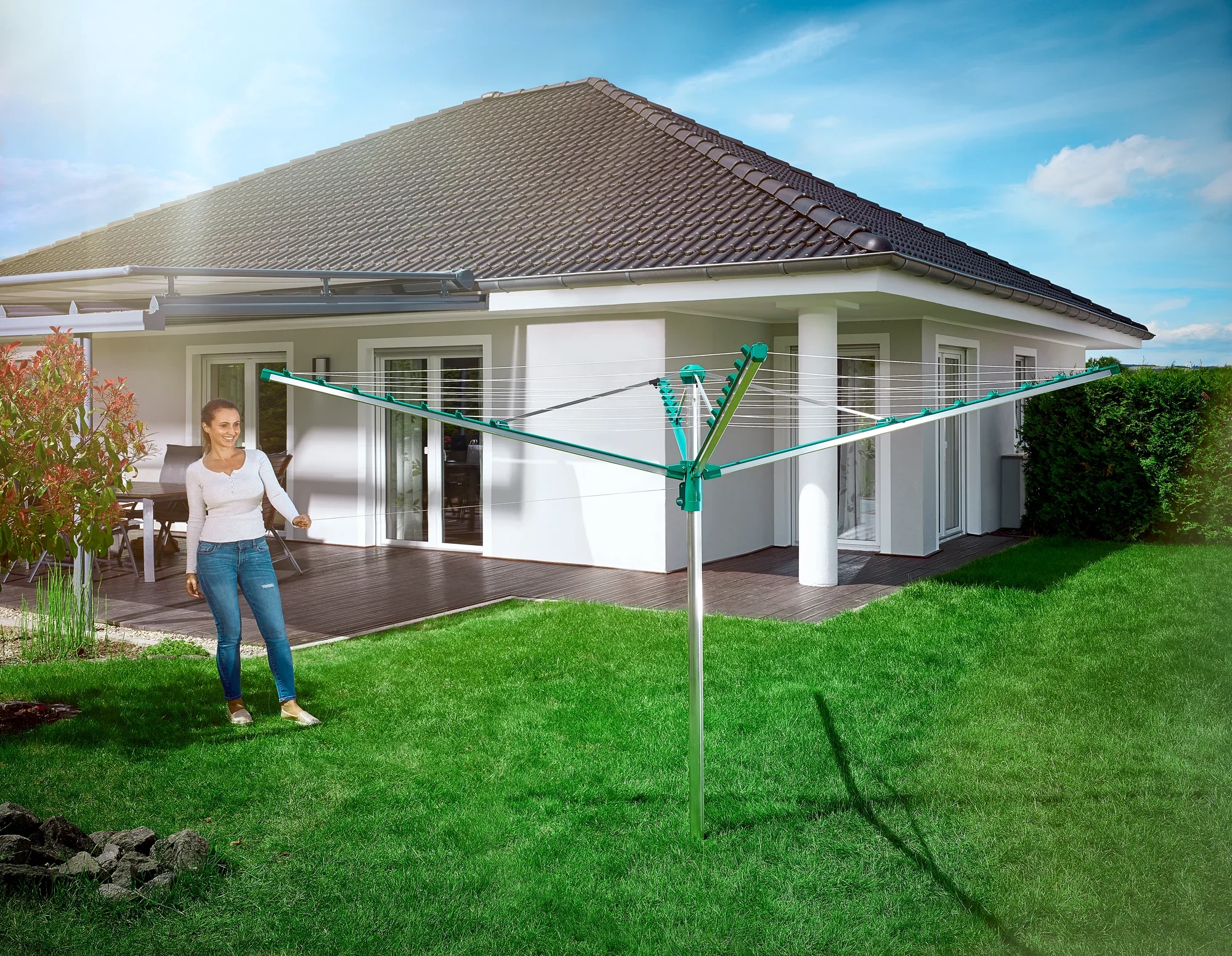 Une femme se tient dans le jardin à côté du séchoir à linge Linomatic 400 Comfort. Le séchoir est ouvert et prêt à accueillir du linge. À l'arrière-plan, on aperçoit une maison individuelle moderne.