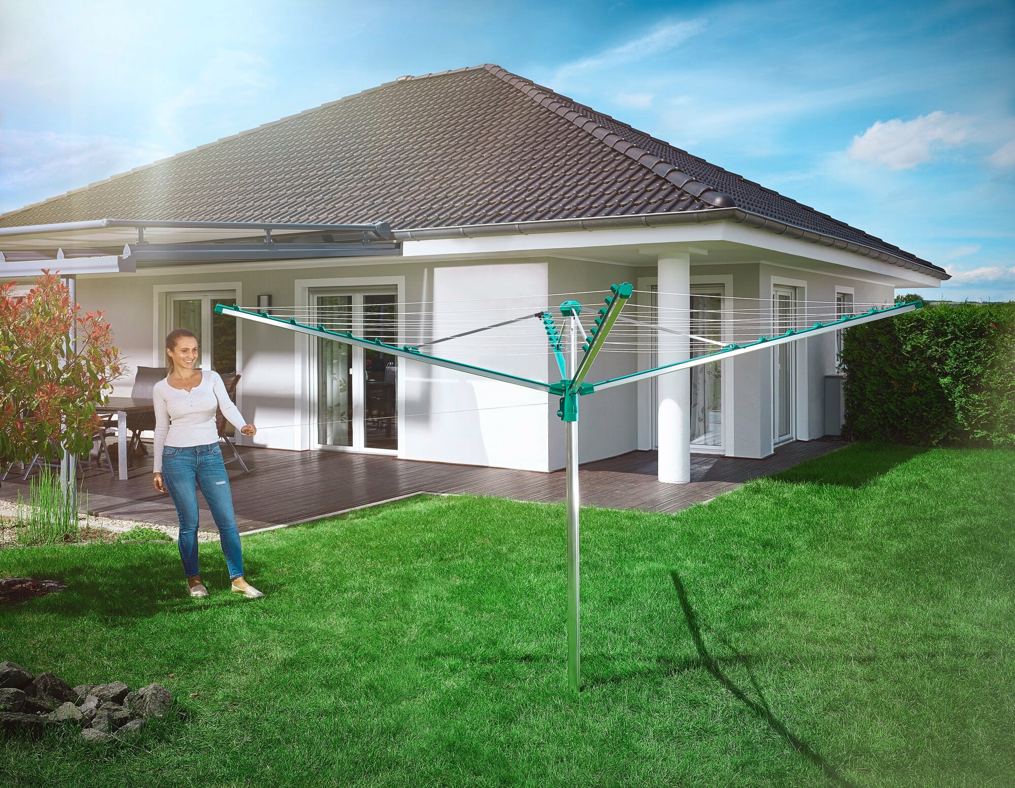 Eine Frau steht im Garten neben der Wäschespinne Linomatic 400 Comfort. Die Wäschespinne ist geöffnet und bereit zum Aufhängen von Wäsche. Im Hintergrund ist ein modernes Einfamilienhaus zu sehen.