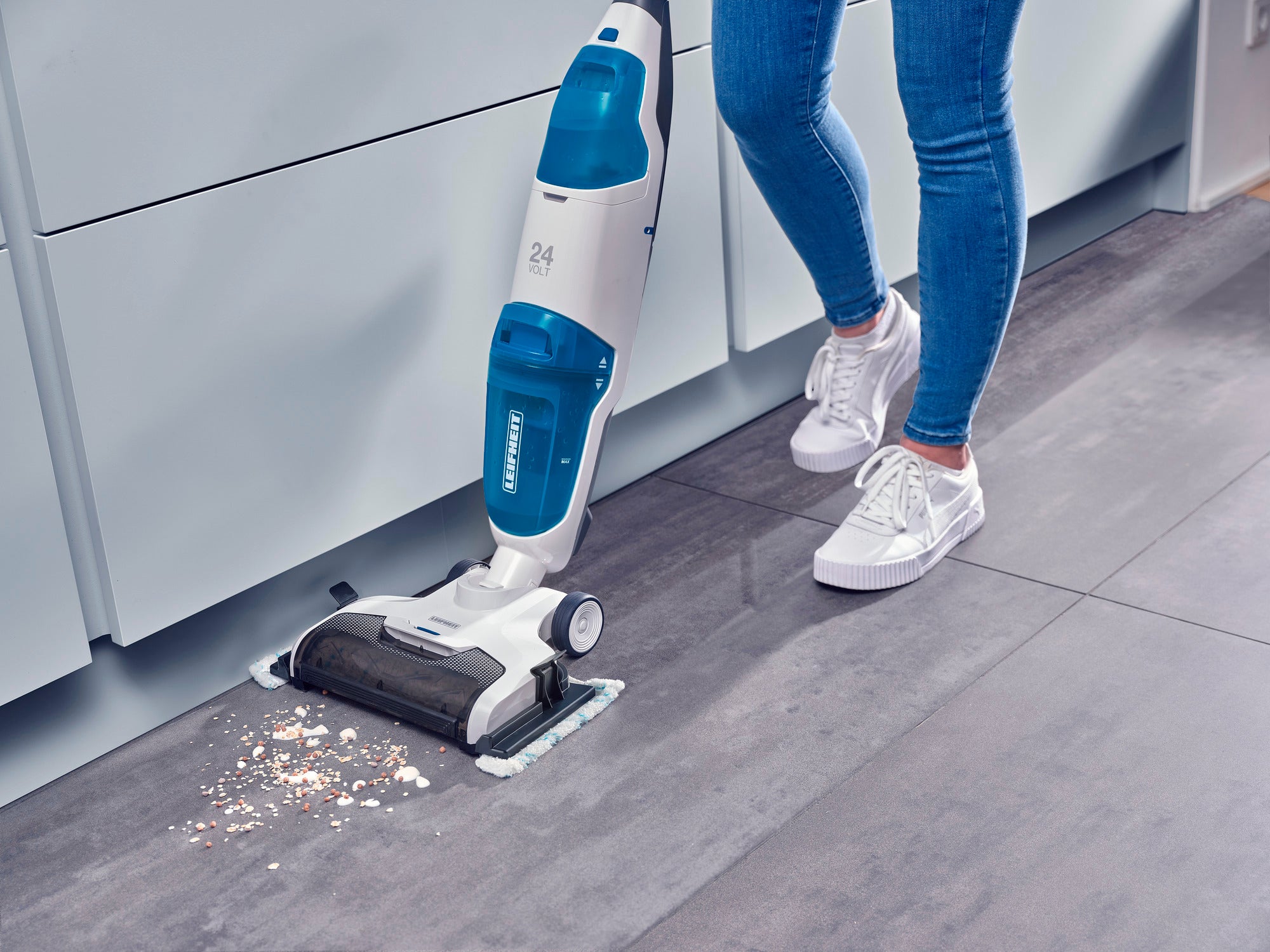 Eine Person verwendet den Regulus Aqua PowerVac Pro, um Krümel von einem Küchenboden zu entfernen. Der Staubsauger ist kabellos und hat ein modernes, blau-weißes Design.